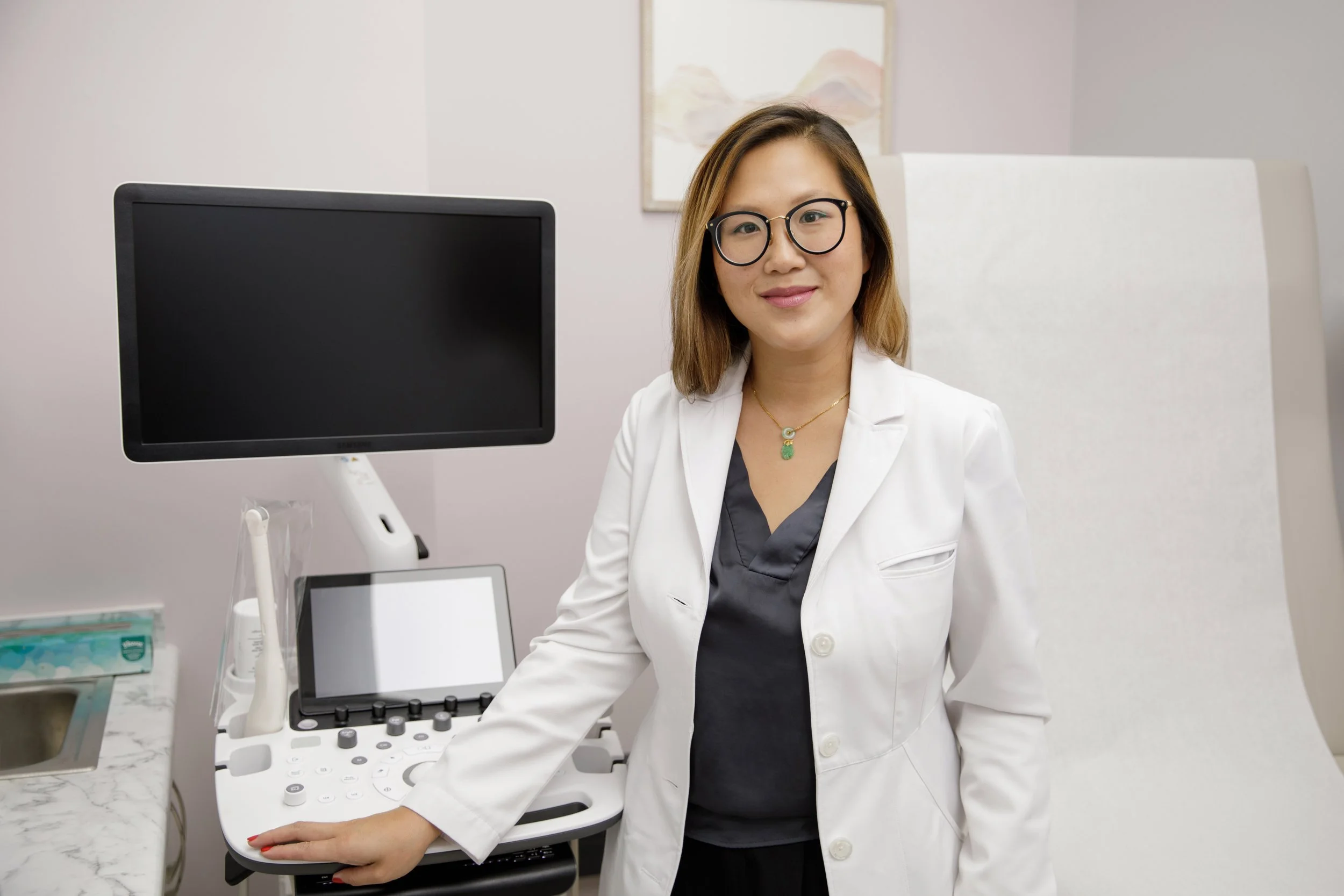 A woman with glasses and a white coat operating a medical ultrasound machine in a clinical setting.