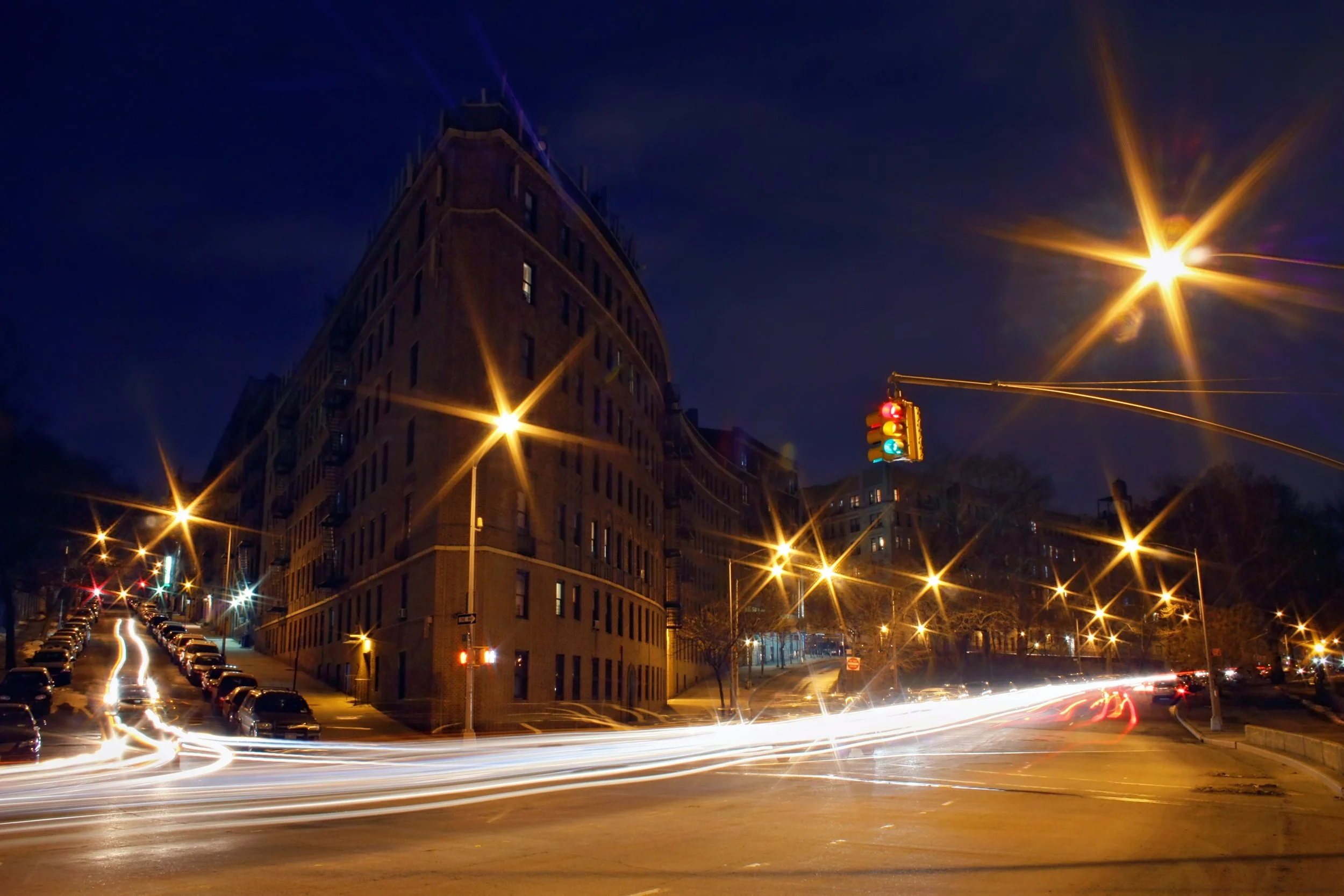 Nighttime city street scene with a tall building and bright streetlights creating starbursts, cars with light trails from long exposure, and a traffic light showing green, yellow, and red lights.