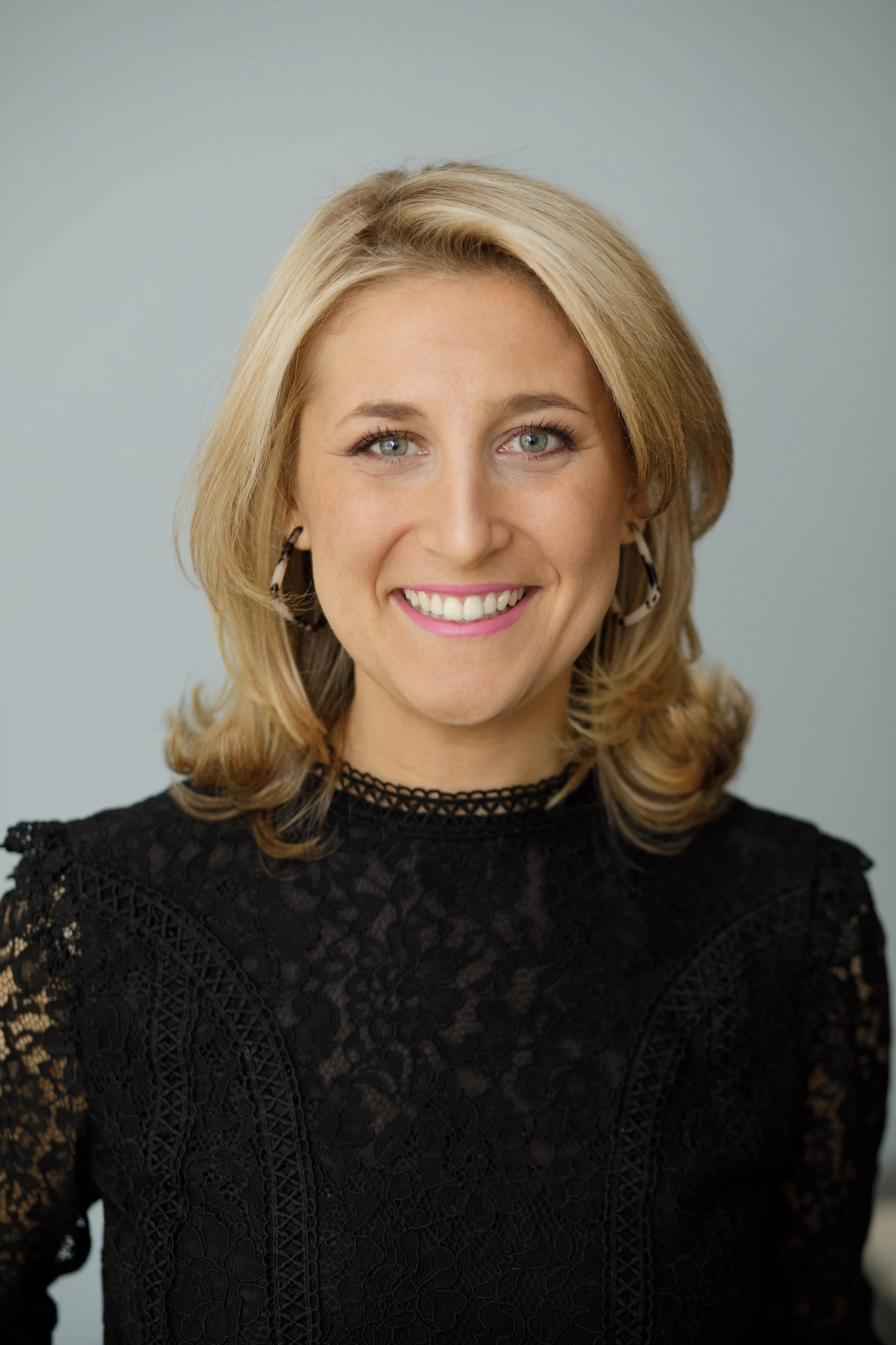 A woman with blonde hair, green eyes, and earrings, smiling while wearing a black lace top.