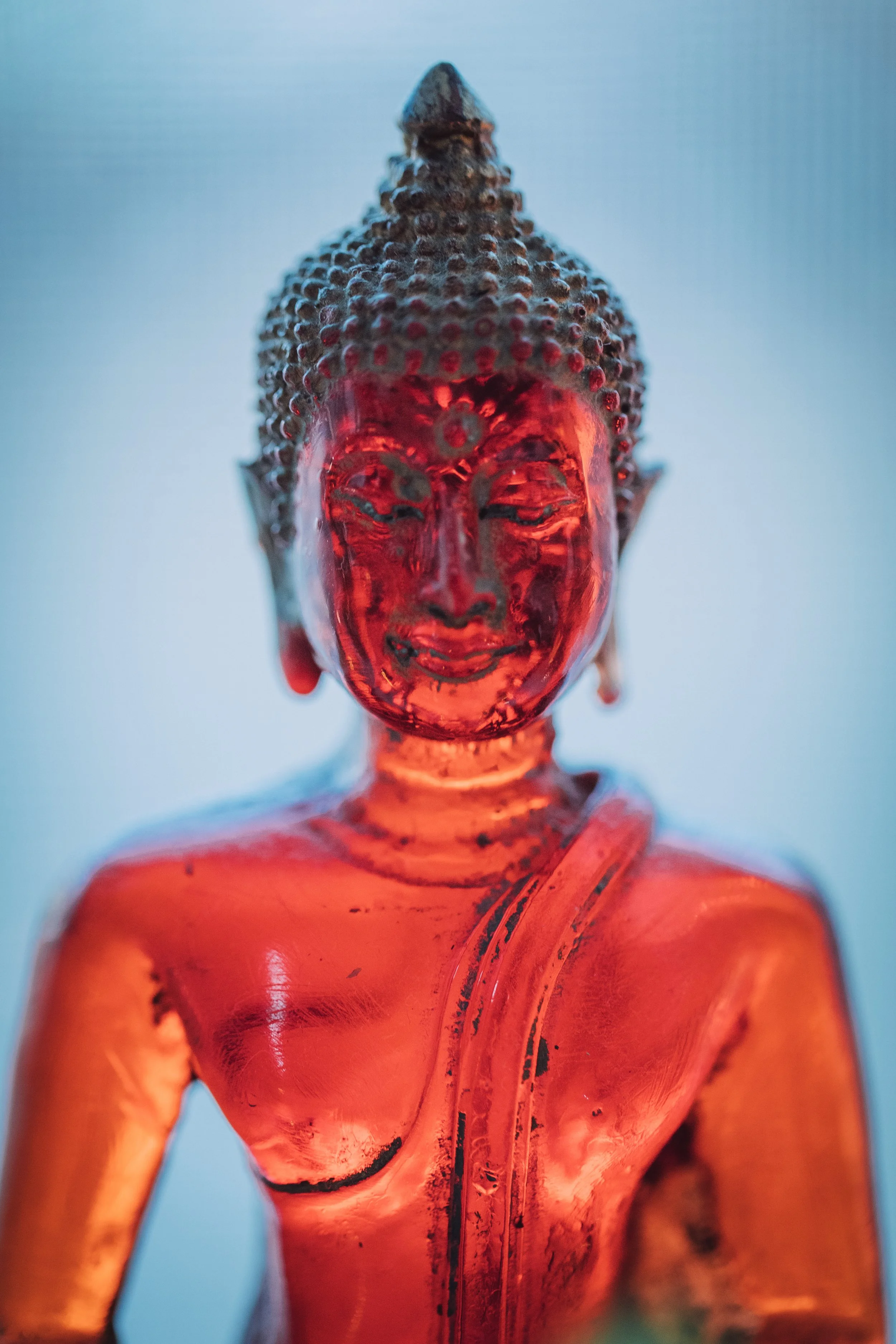 A close-up of a red-colored Buddha statue with a smiling face, detailed hair, and an arm crossed over the chest, against a light background.