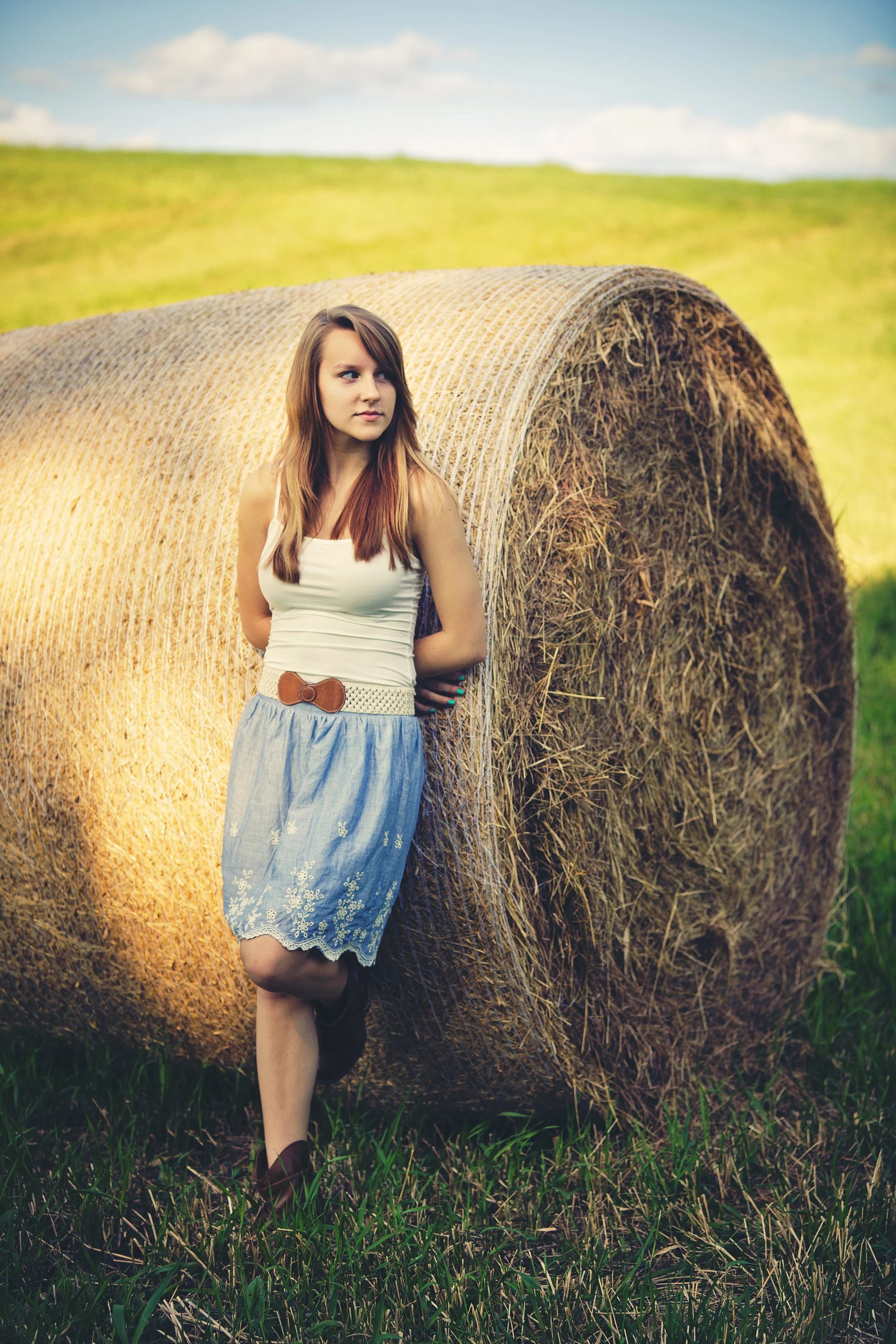 A young woman with long, light brown hair wearing a white tank top, a blue skirt, and dark brown boots leaning against a large hay bale in a field with green grass and a blue sky with clouds in the background.