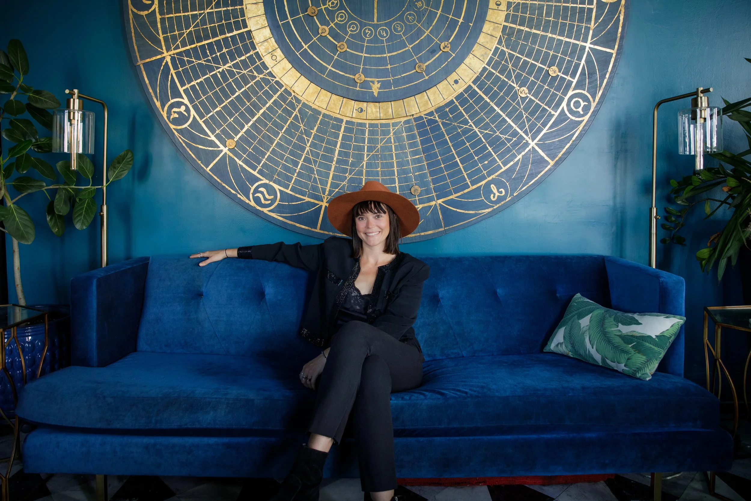 A woman with dark hair wearing a brown hat and black outfit sitting on a blue velvet sofa in front of a large, circular, gold and blue celestial-themed wall art. She is smiling and has her arm resting on the back of the sofa. There are green plants a