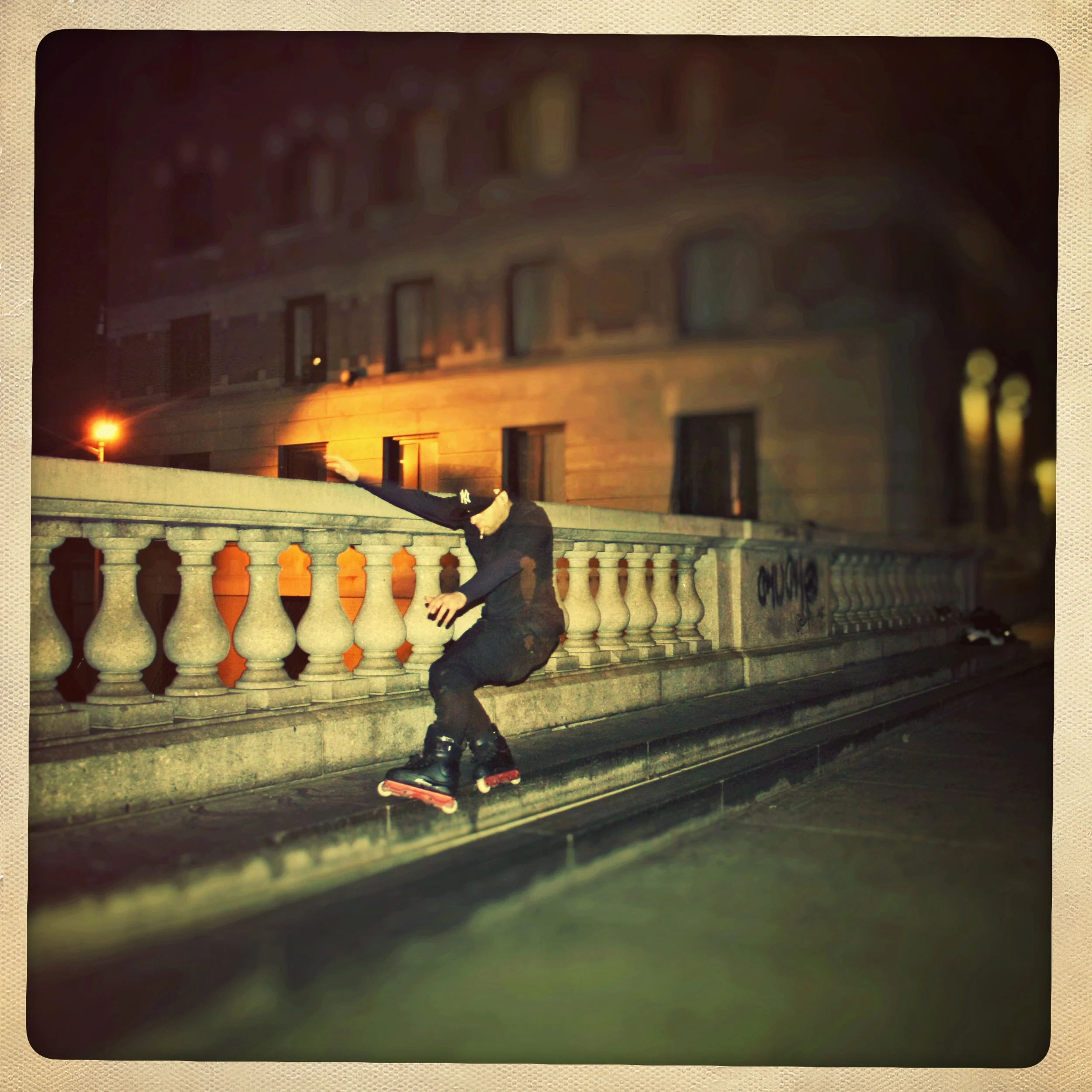 A person standing on rollerblades on a balcony at night, holding onto the railing. There is a building with lit windows in the background and streetlights below.
