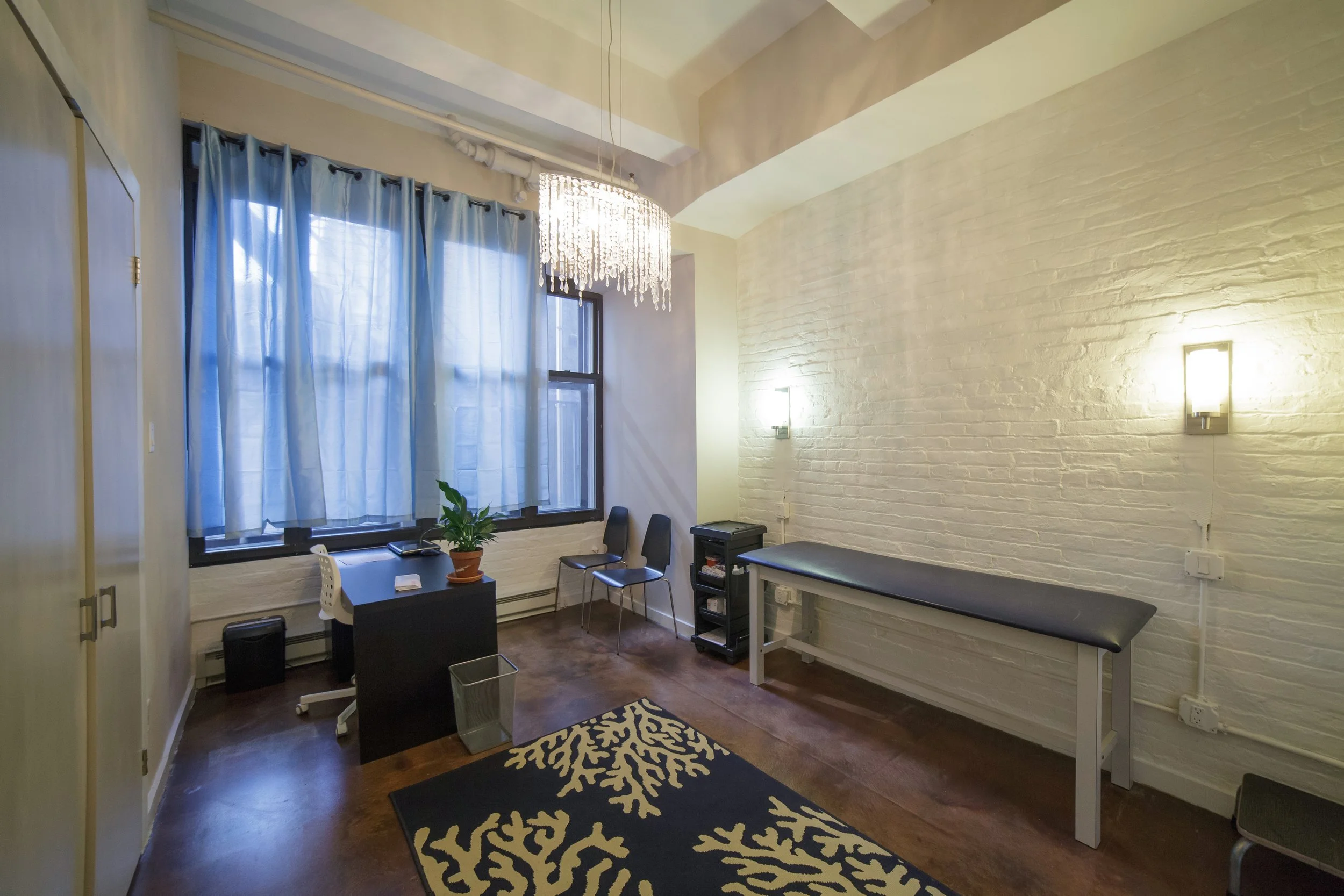 Office room with blue curtains, white brick walls, a chandelier, a desk with a plant, chairs, and a black and white patterned rug on a wooden floor.