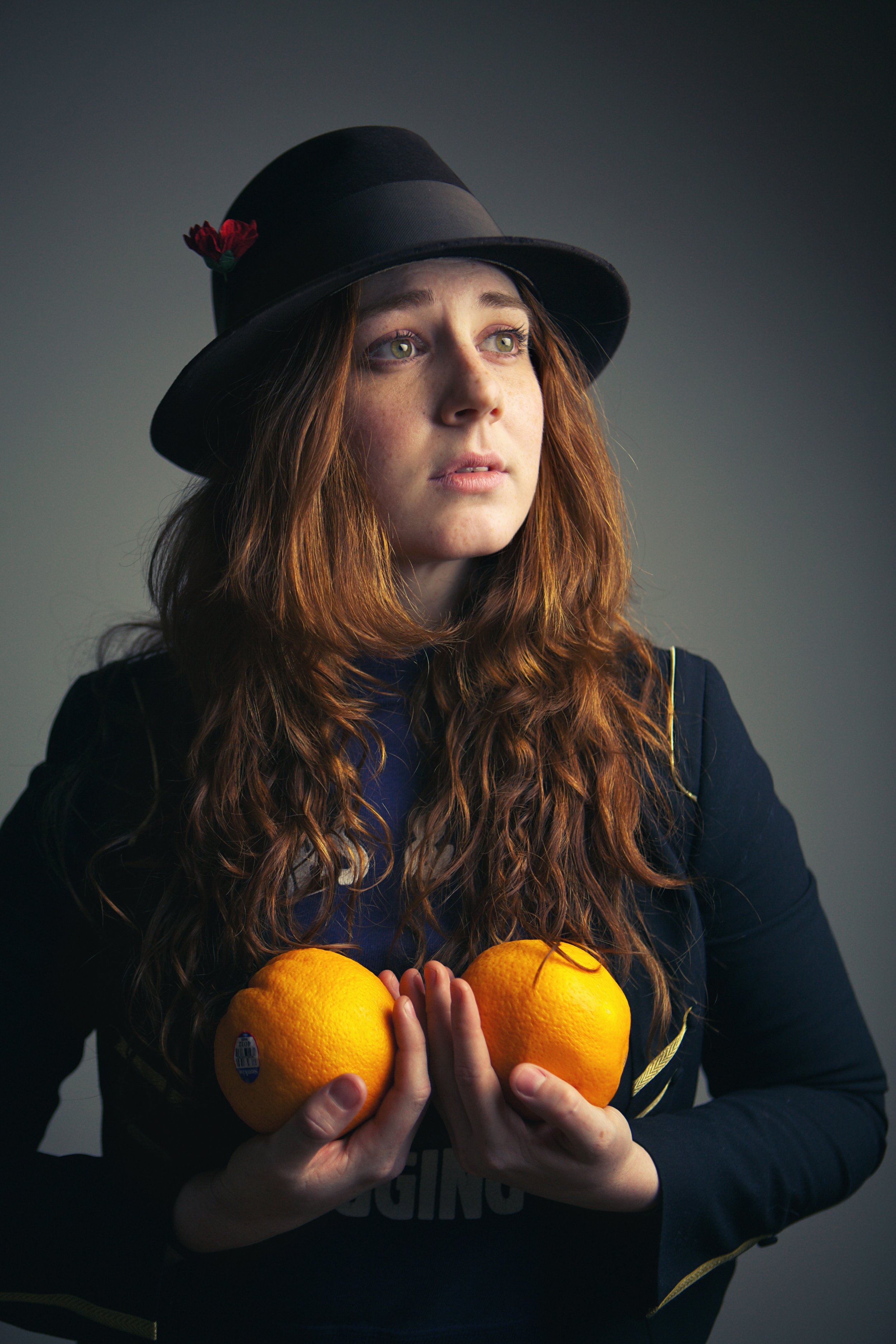 A young woman with long red hair, wearing a black hat with a small red flower, holds three large oranges in front of her. She wears a dark jacket with yellow accents and looks pensively to the side.