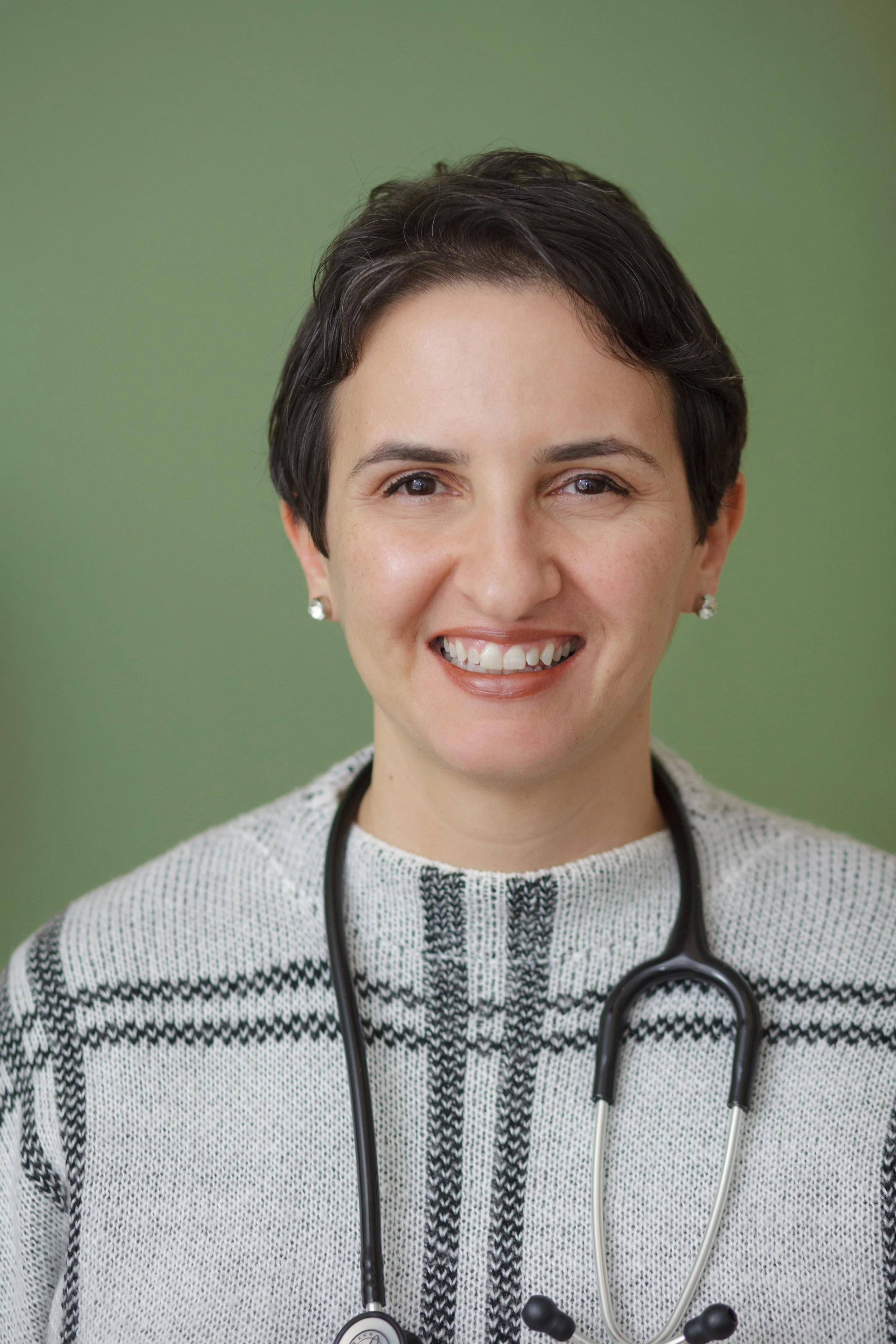 Close-up of a smiling female doctor or nurse with short dark hair, wearing a white and black sweater, earrings, and a stethoscope around her neck, against a green background.