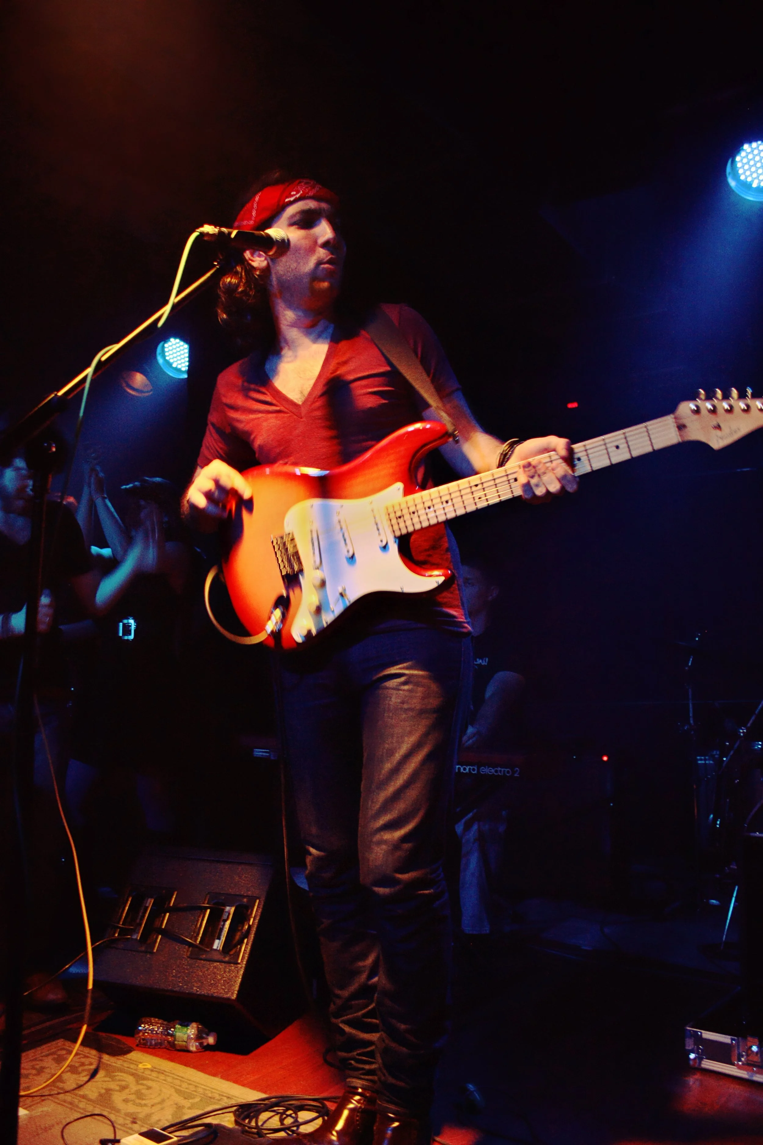 A person playing an electric guitar on stage with stage lighting and musical equipment around.