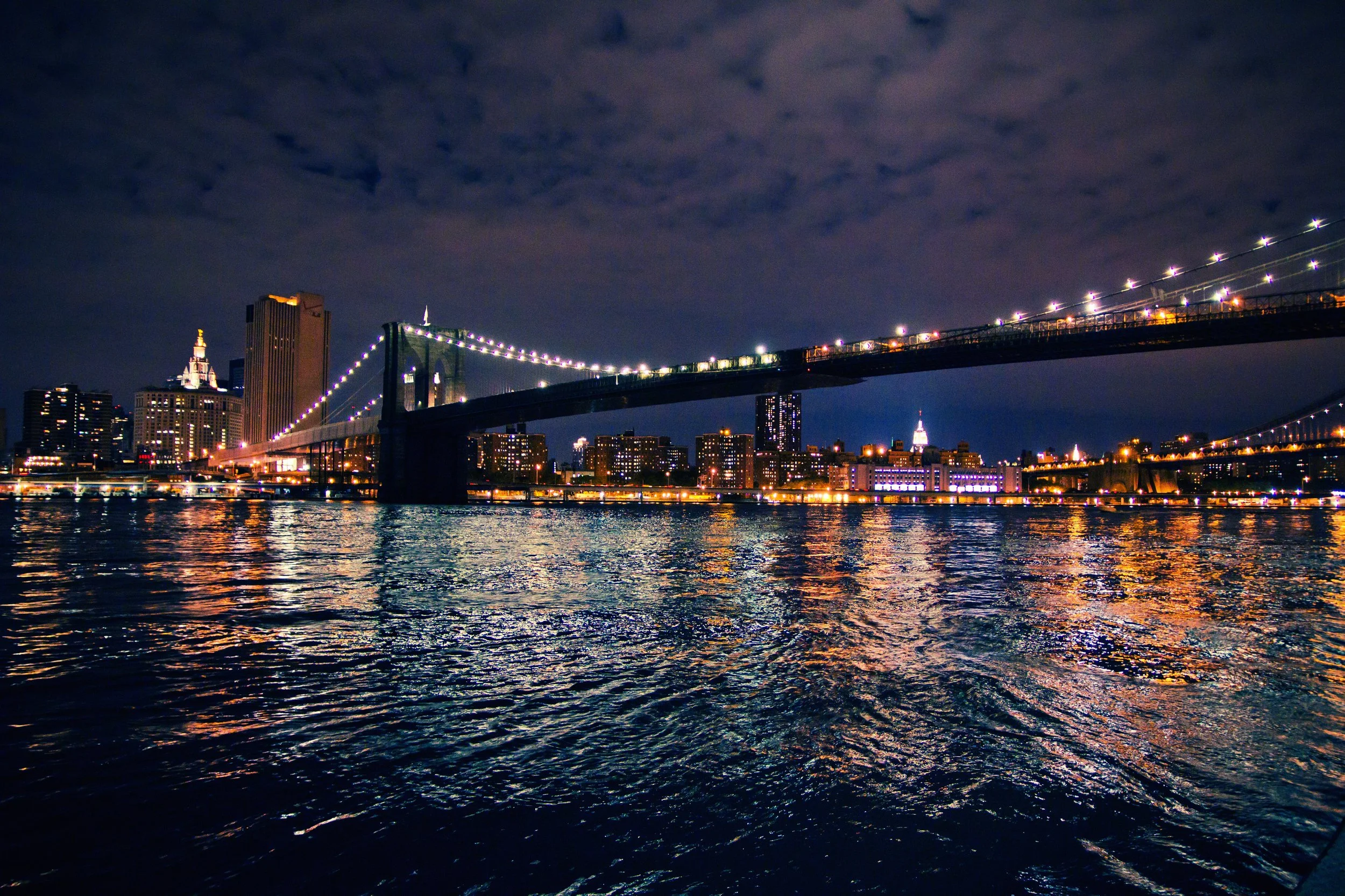 Night view of a city skyline with illuminated buildings and a bridge over water, with clouds in the sky.