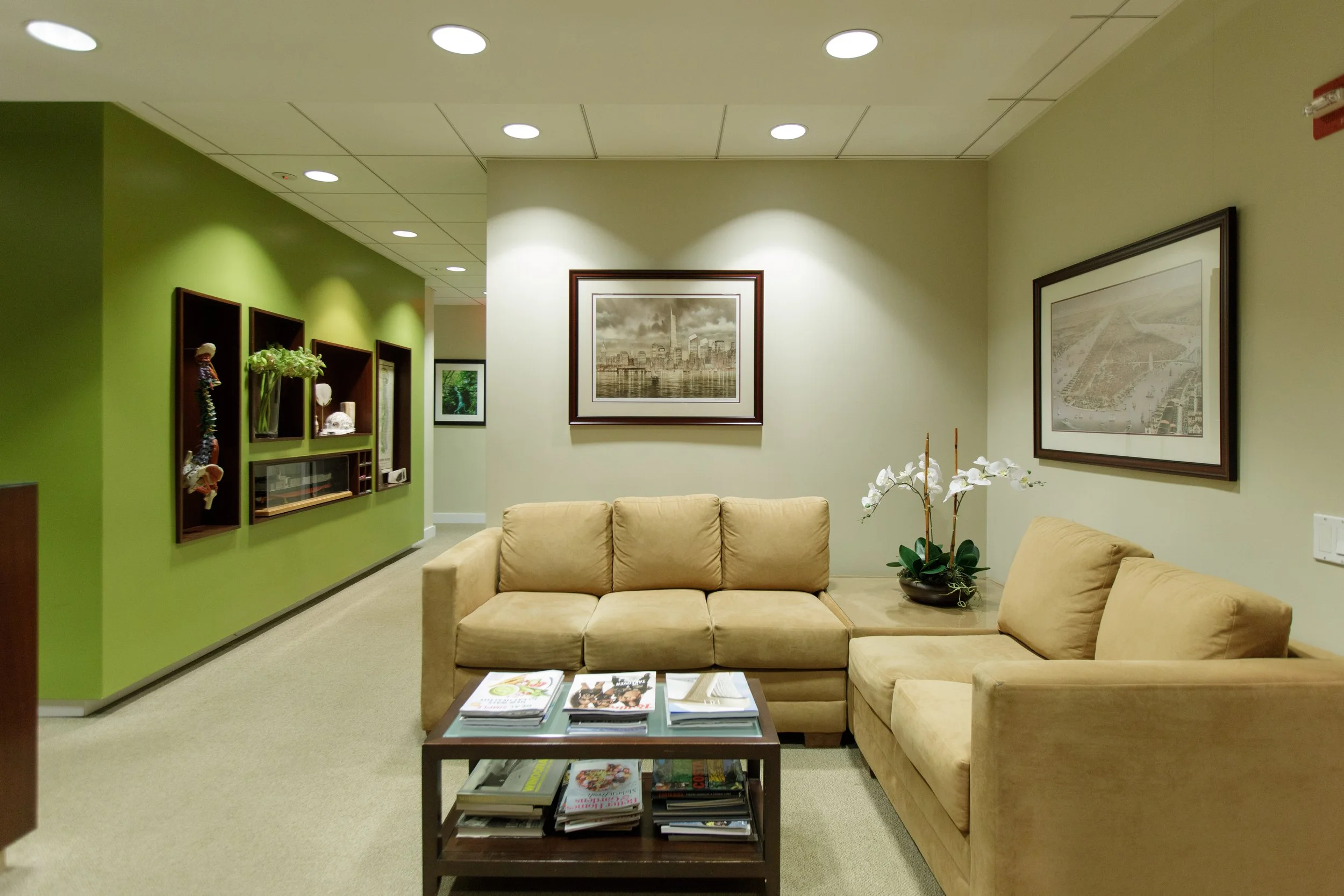A beige sectional sofa in a waiting room with a coffee table full of magazines and brochures, decorated with a white orchid plant, against a light-colored wall with framed pictures and a green accent wall with display shelves.