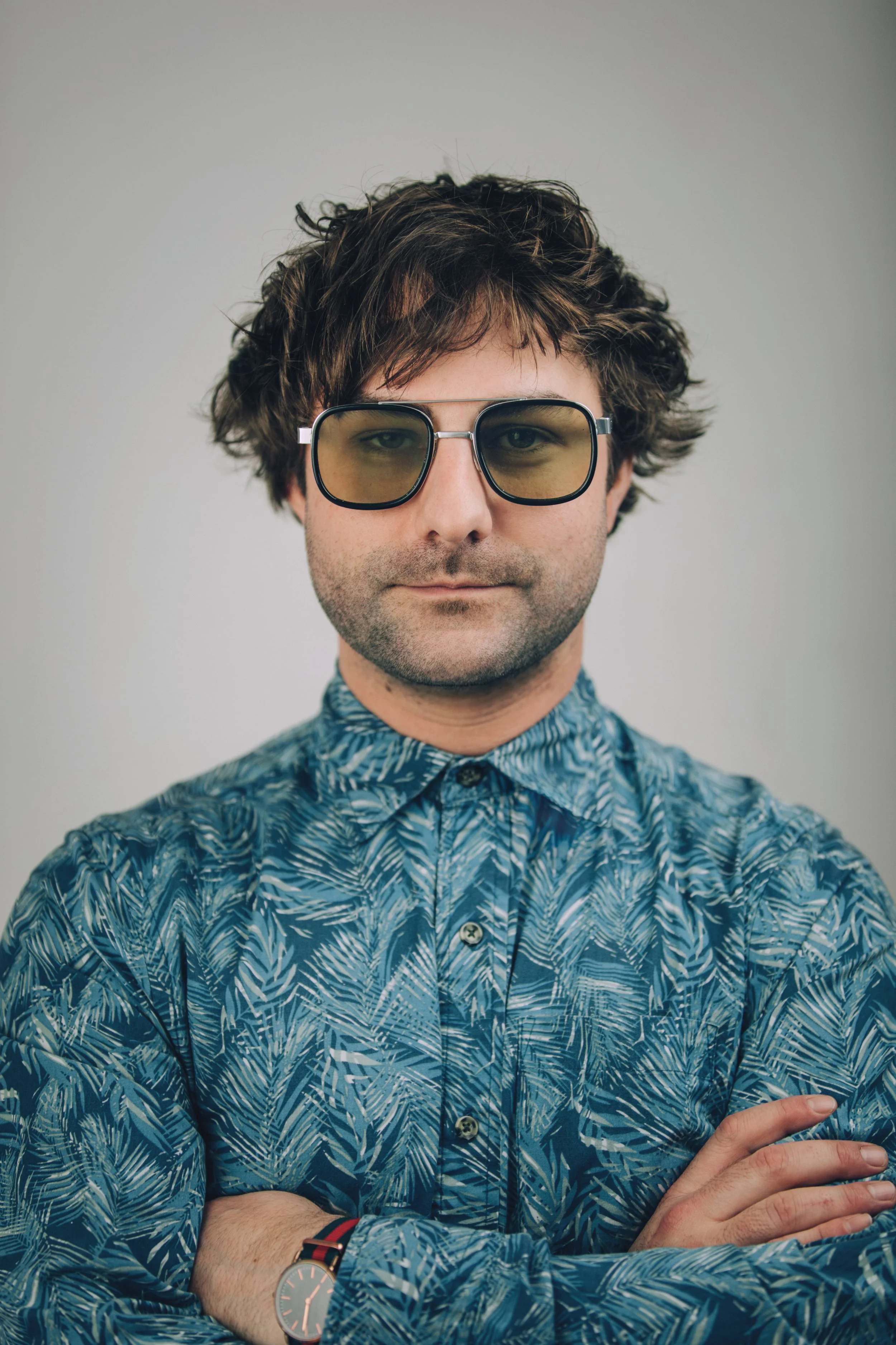 Man with curly brown hair wearing large yellow-tinted sunglasses and a blue patterned shirt, crossing his arms, with a wristwatch visible on his left wrist.