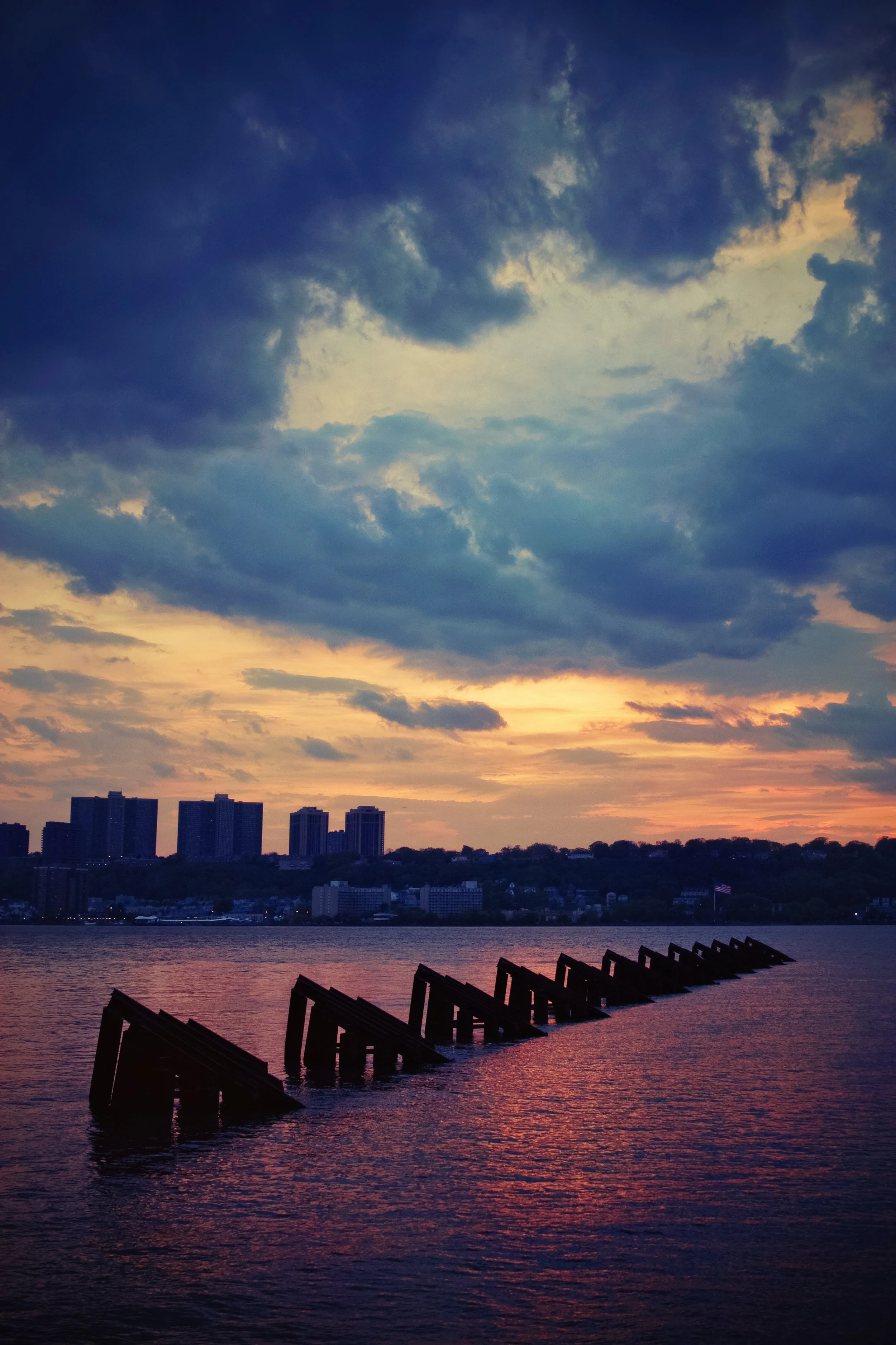 Sunset over a city skyline with high-rise buildings and a river in the foreground, with silhouettes of structures in the water and clouds in the sky.