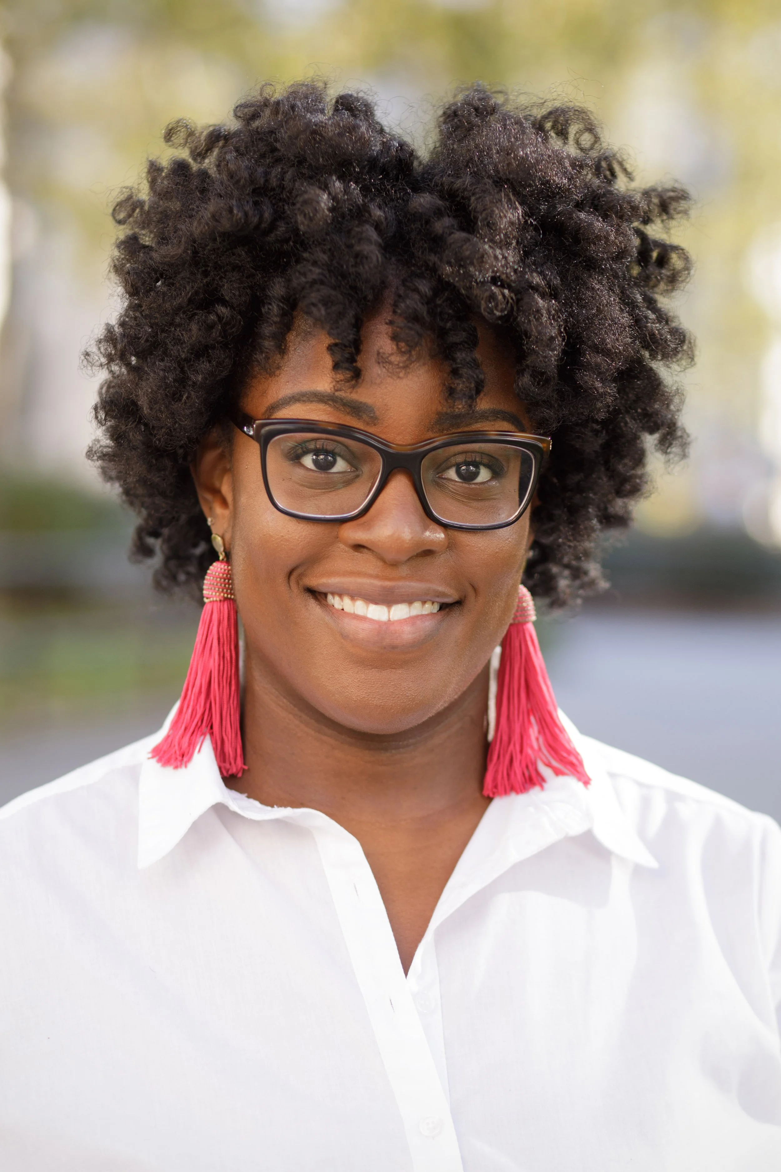 Close-up of a woman with curly black hair wearing glasses, earrings, a white shirt, and pink tassel earrings outdoors.