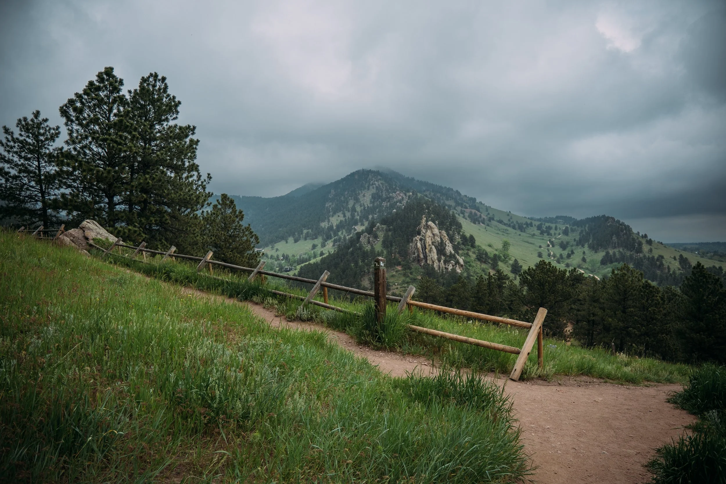 A mountainous landscape with a dirt trail, a wooden fence, trees, and cloudy sky.