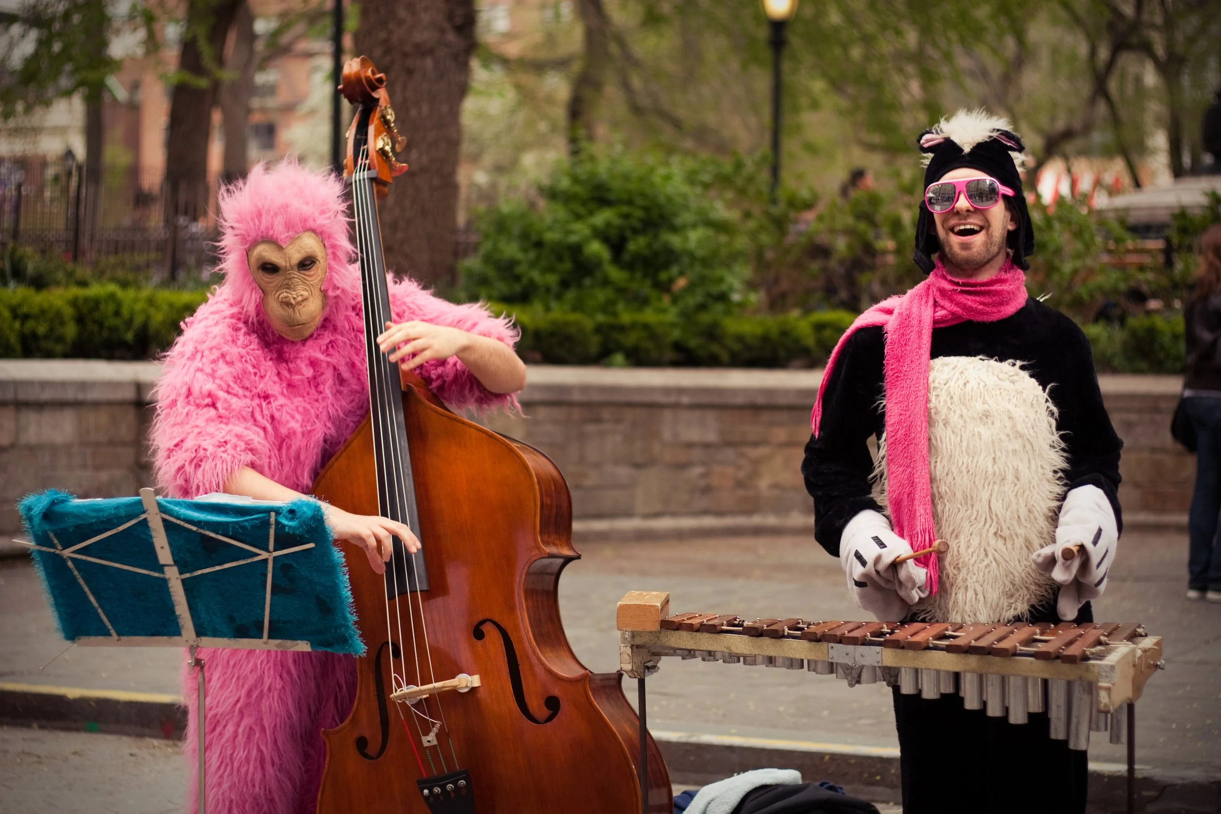 Person wearing a pink furry costume playing a double bass and a music stand with blue furry cover, another person in a black and white furry costume with pink sunglasses and a pink scarf, both outdoors in a park with trees and fencing in the backgrou