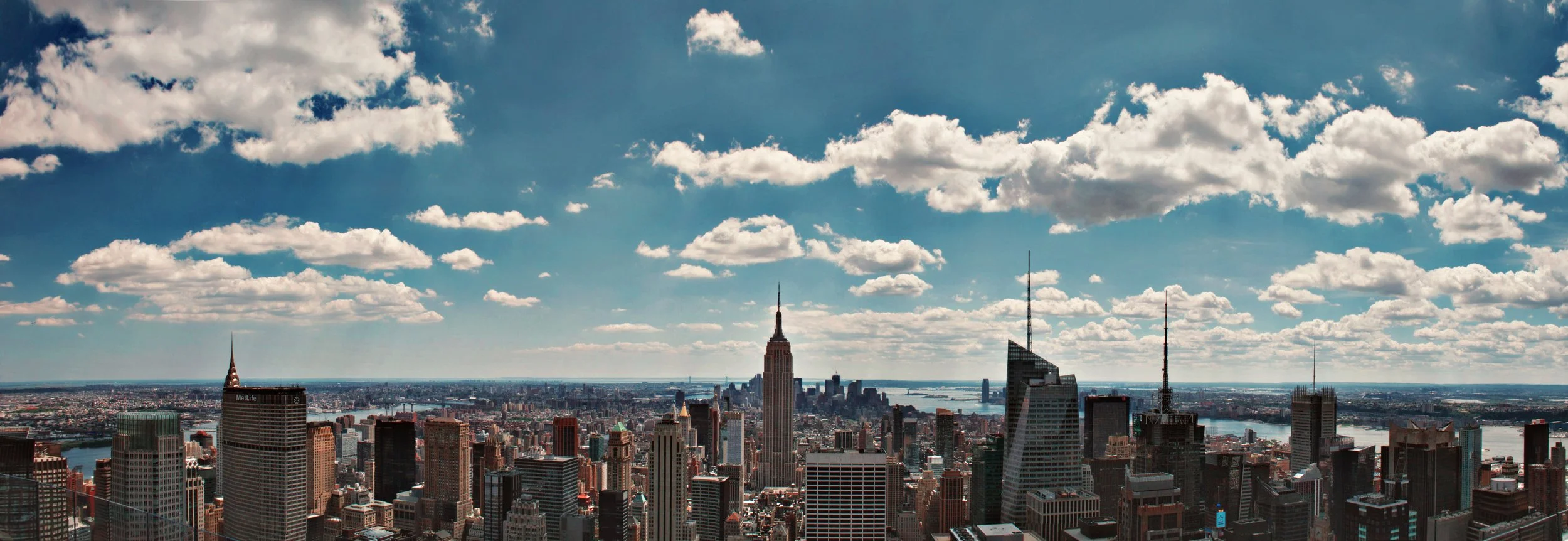A panoramic view of Manhattan skyline with tall skyscrapers, the Empire State Building in the center, and a river in the background under a partly cloudy sky.