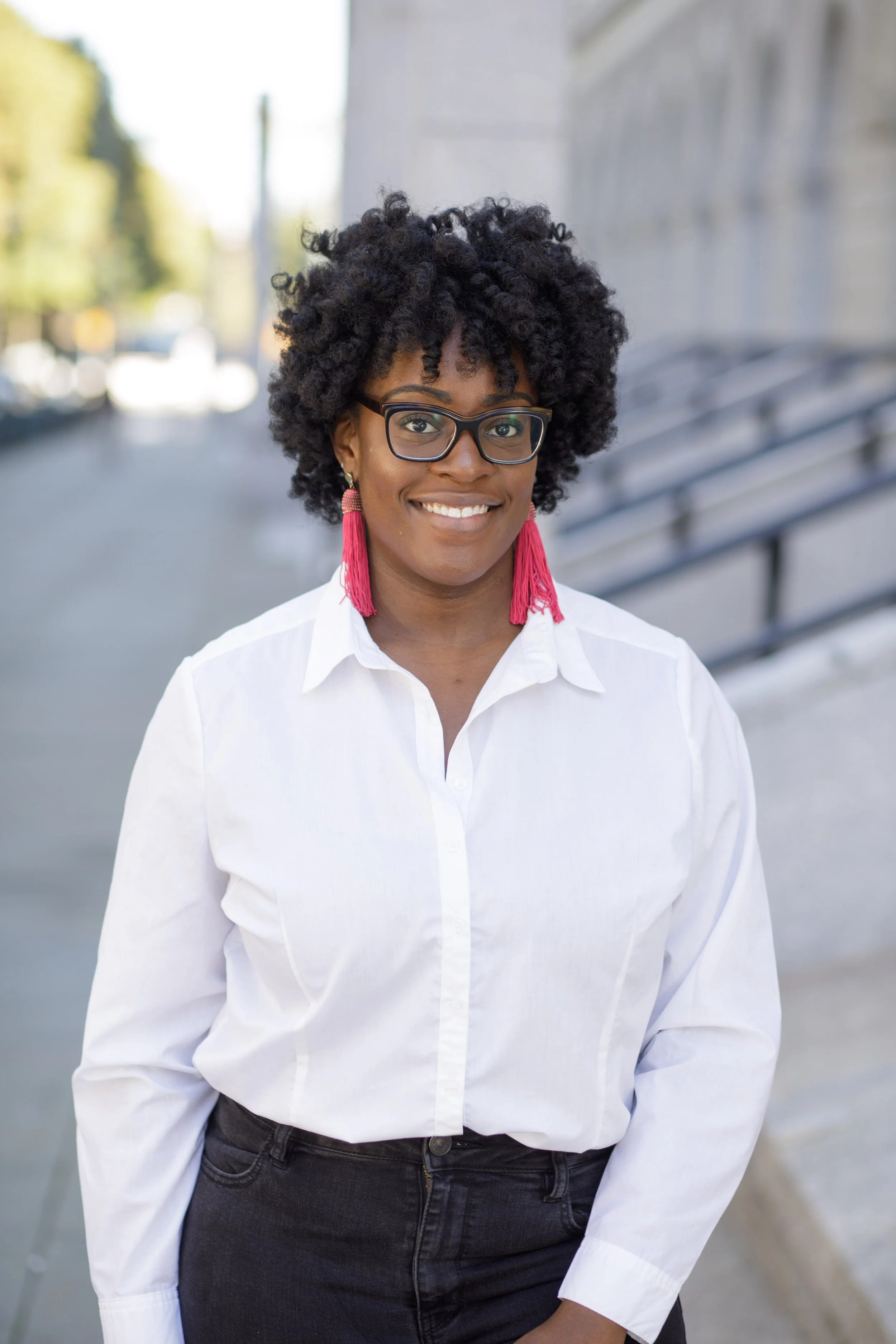 A woman with curly black hair, glasses, and pink tassel earrings smiling outdoors. She is wearing a white button-up shirt and black pants, standing near a bench against a blurred urban background.