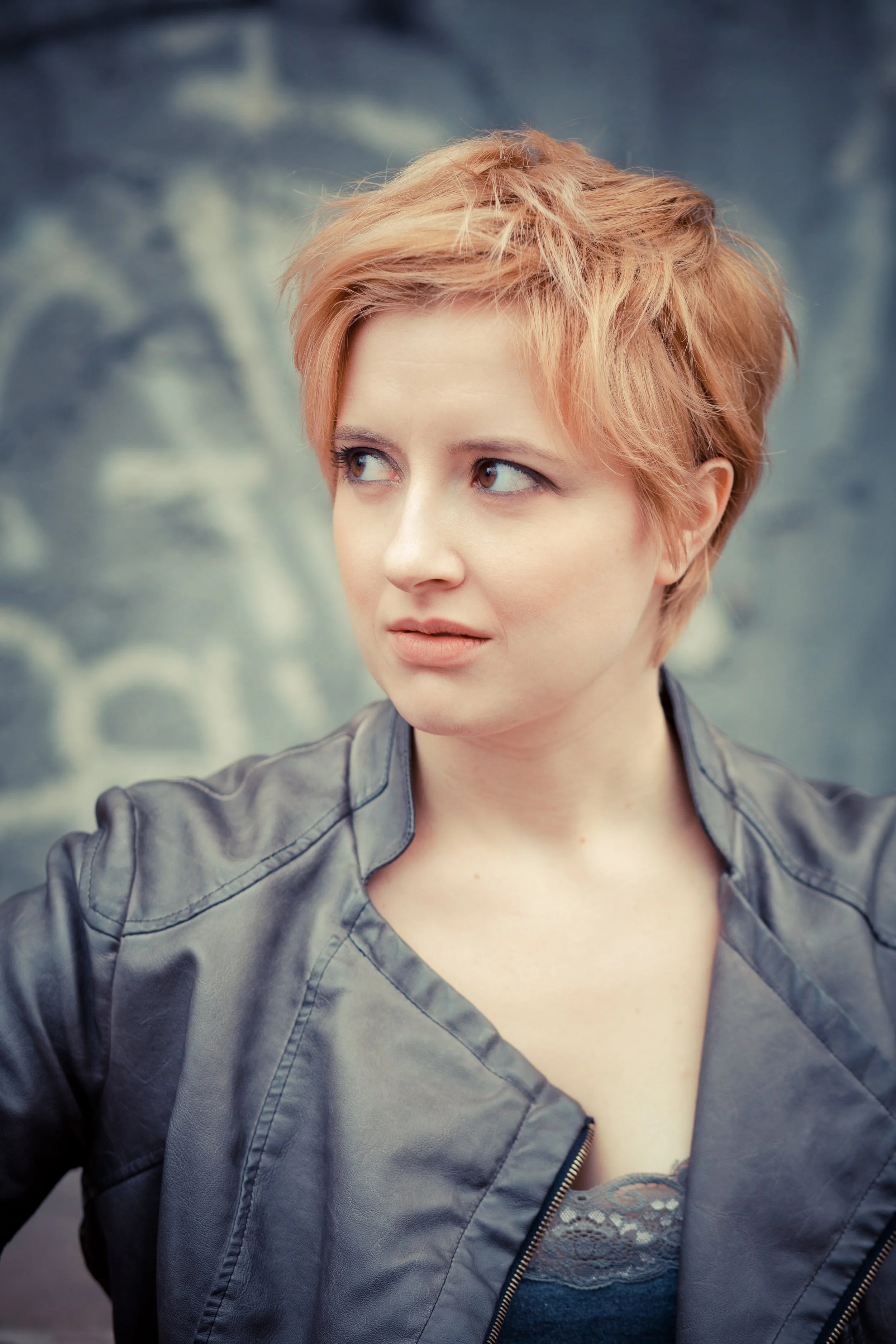 A woman with short orange hair wearing a gray leather jacket, looking to her right, against a blurred outdoor background.