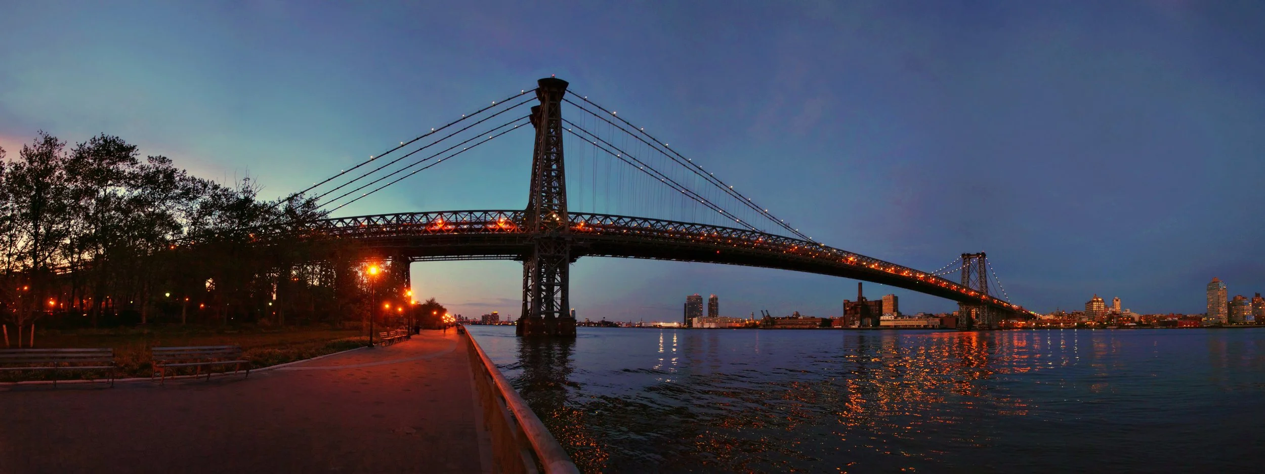 A view of a large suspension bridge illuminated at dusk, spanning over a body of water with city skyline in the background.