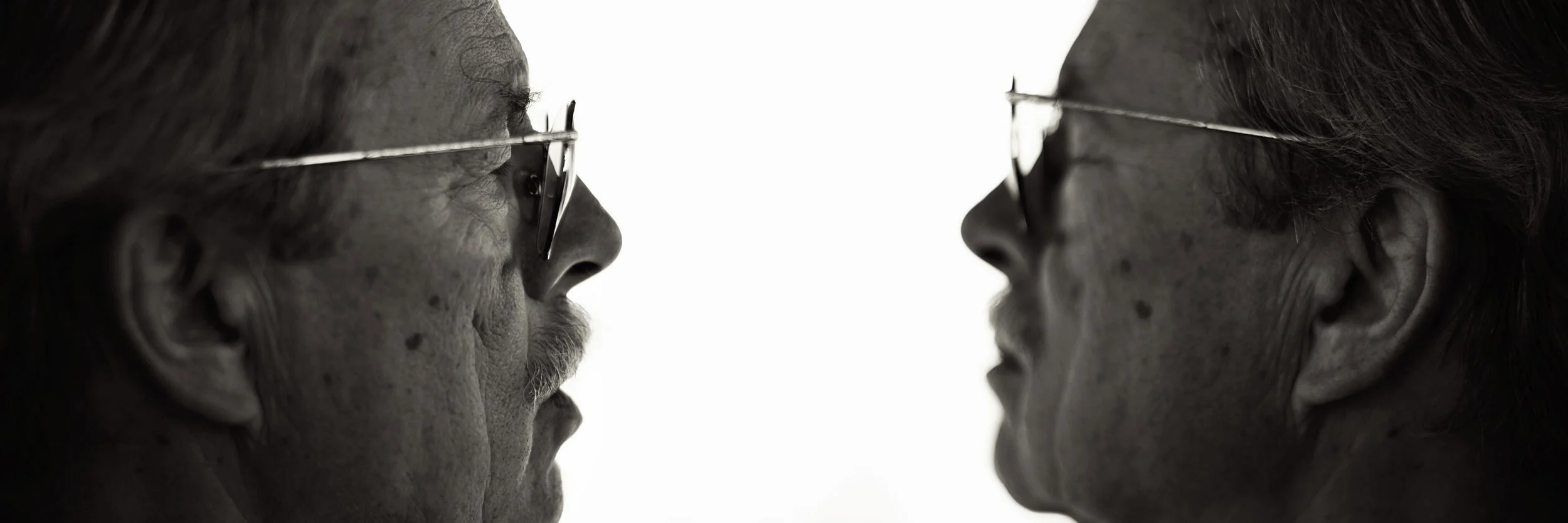 Close-up of two elderly women facing each other with their noses nearly touching, both wearing glasses.