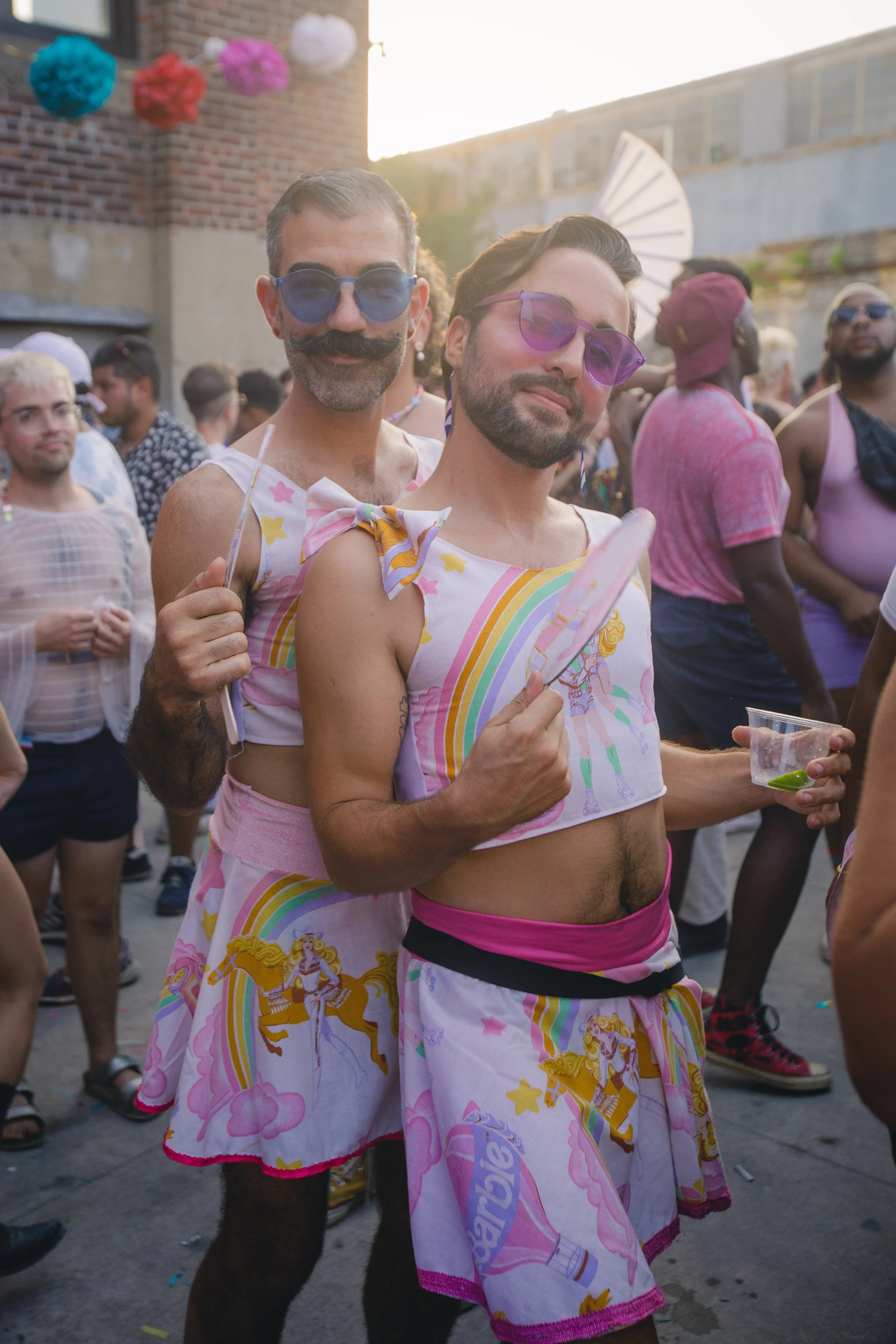 Two men dressed in matching colorful rainbow and unicorn-themed outfits at a pride parade, holding drinks and standing among a crowd of diverse people.