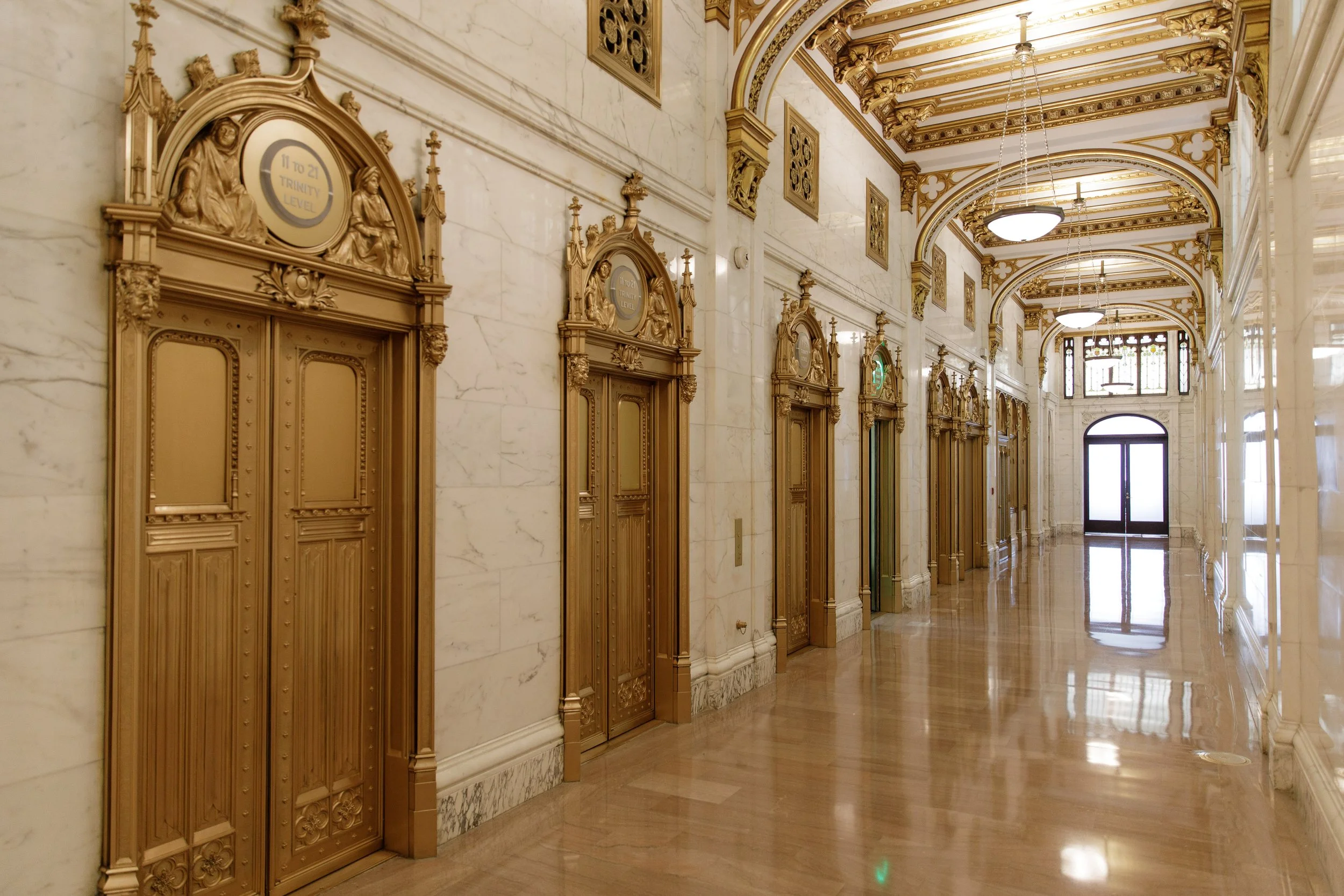 Elegant hallway with marble walls and floors, ornate gold-colored elevator doors with intricate carvings, decorative gold and white ceiling with chandeliers, and large windows at the end of the hallway.