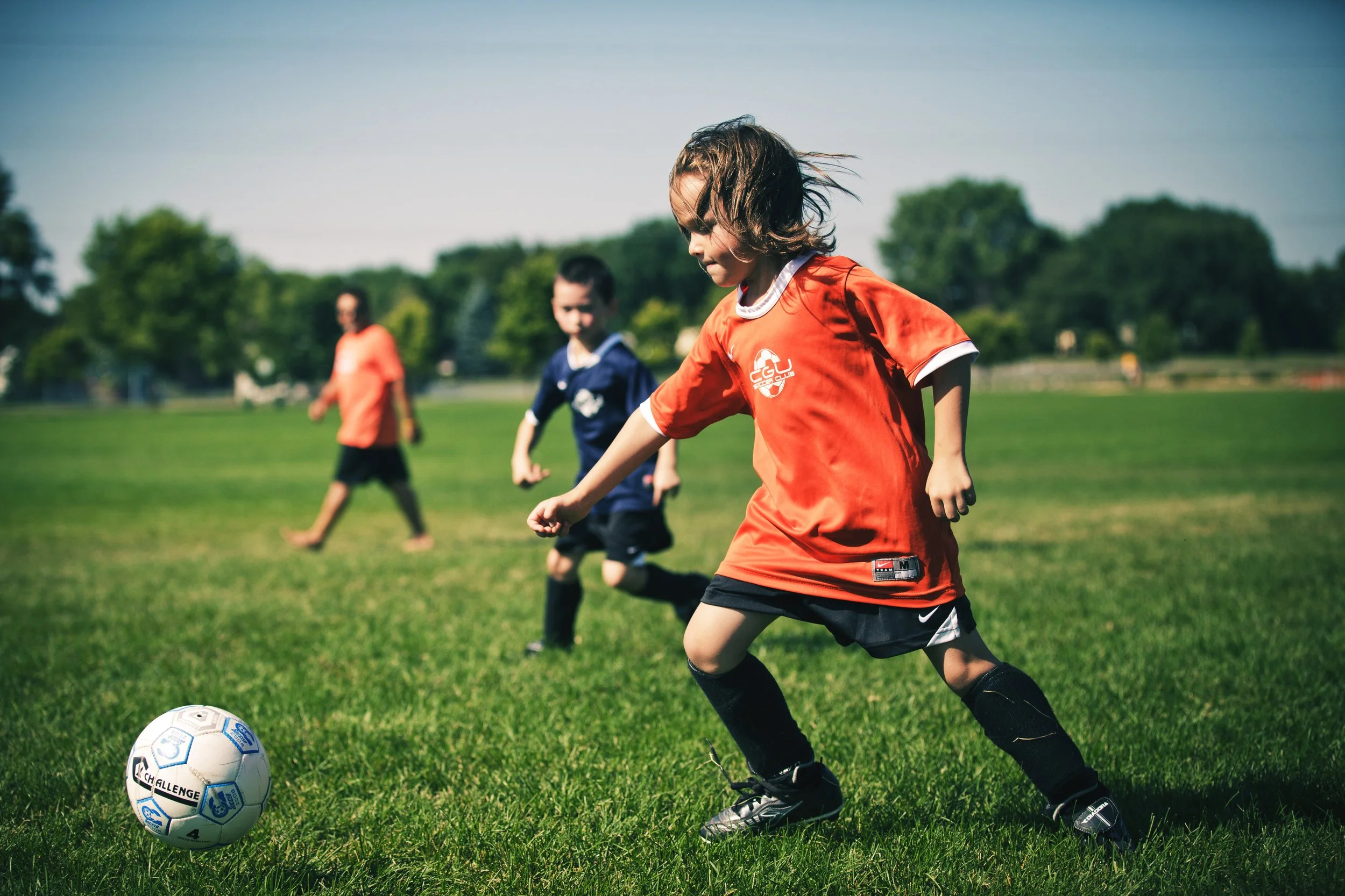 Children playing soccer on a green field, wearing sports uniforms, with one child in focus kicking a soccer ball.