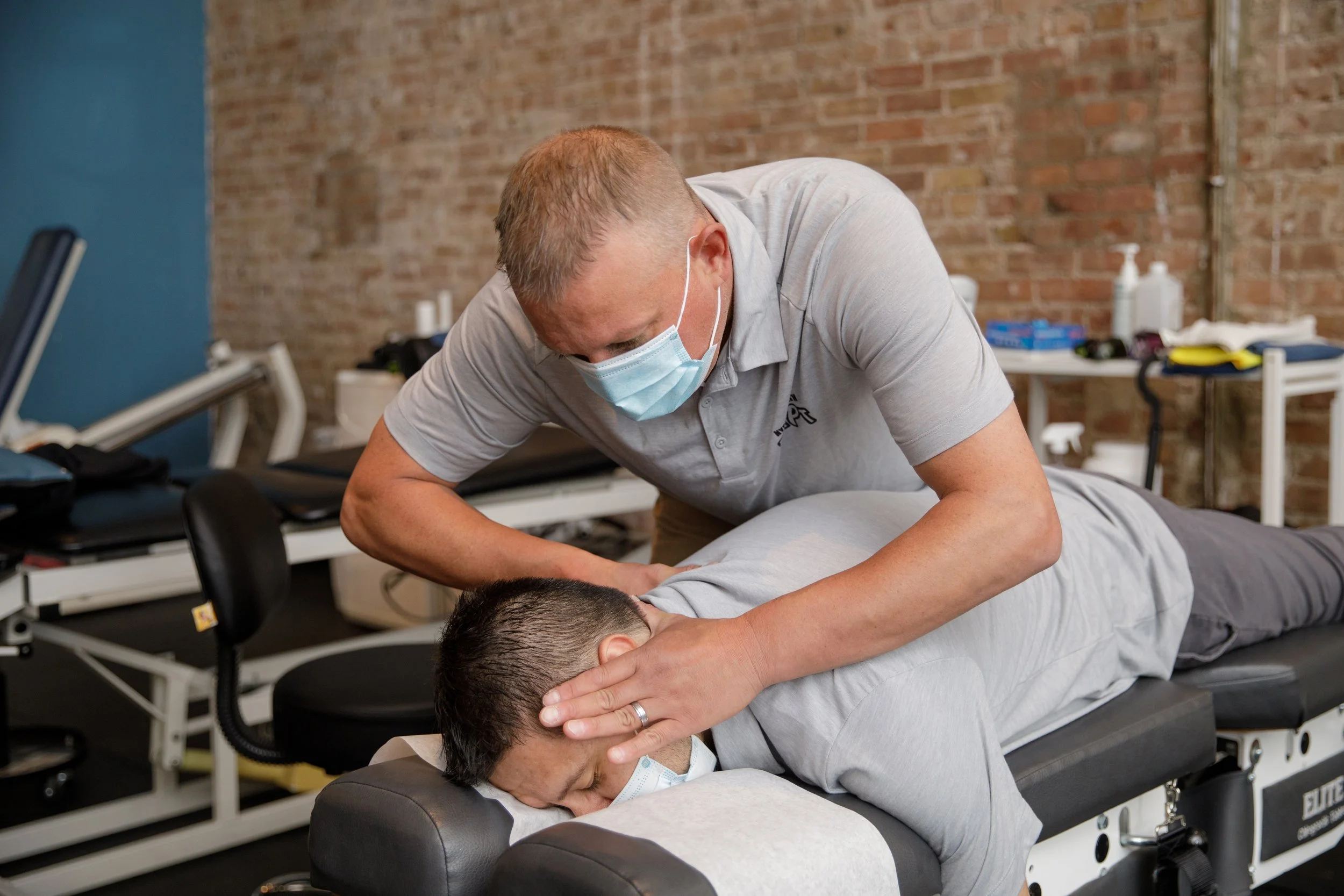A chiropractor wearing a face mask adjusting a patient's neck during a therapy session in a clinic with brick walls.