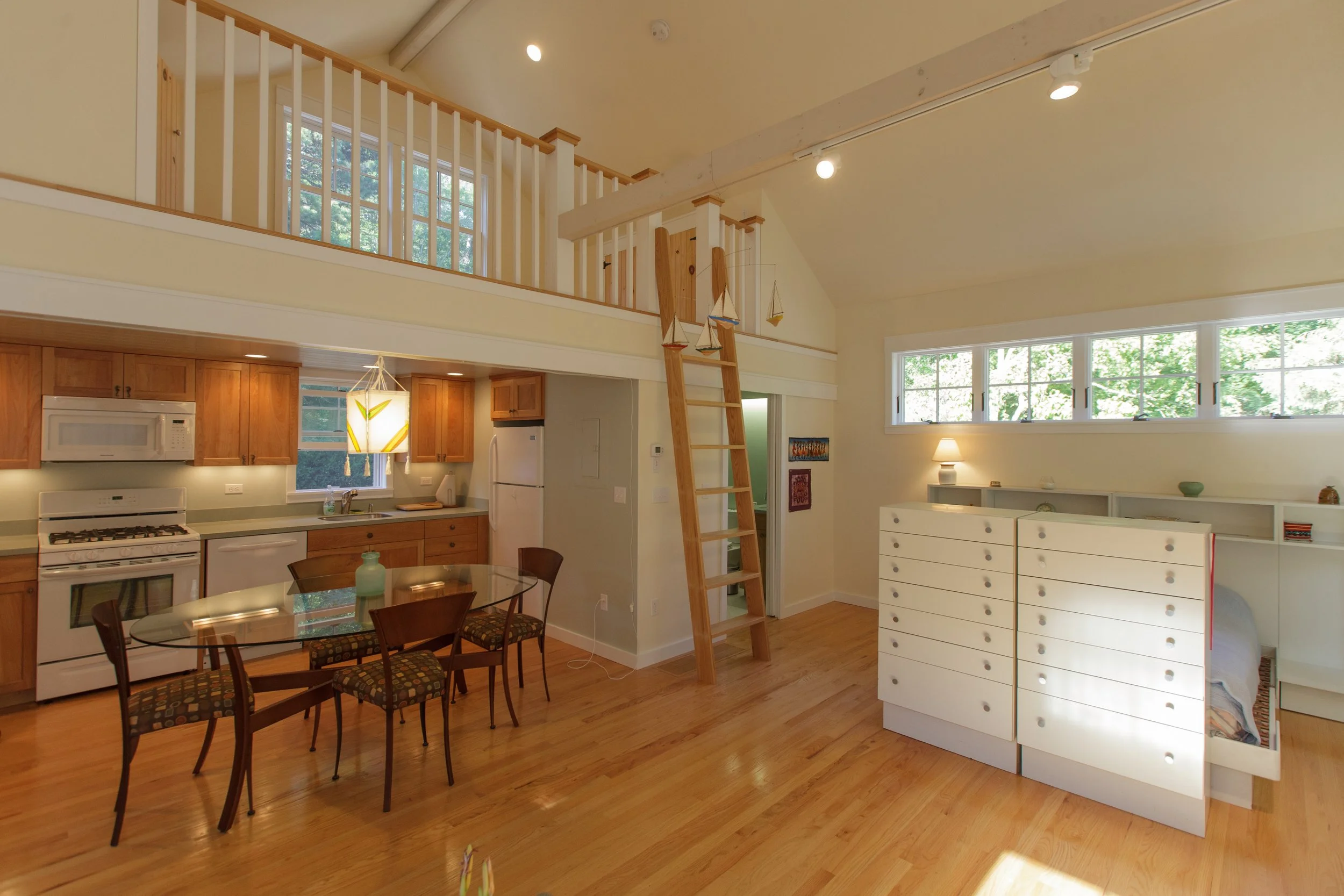 Interior of a living space with a kitchen and dining area, featuring wooden floors, a kitchen with white appliances and wooden cabinets, a glass-top dining table with four chairs, and a loft area with a wooden ladder, decorated with sailboat ornament