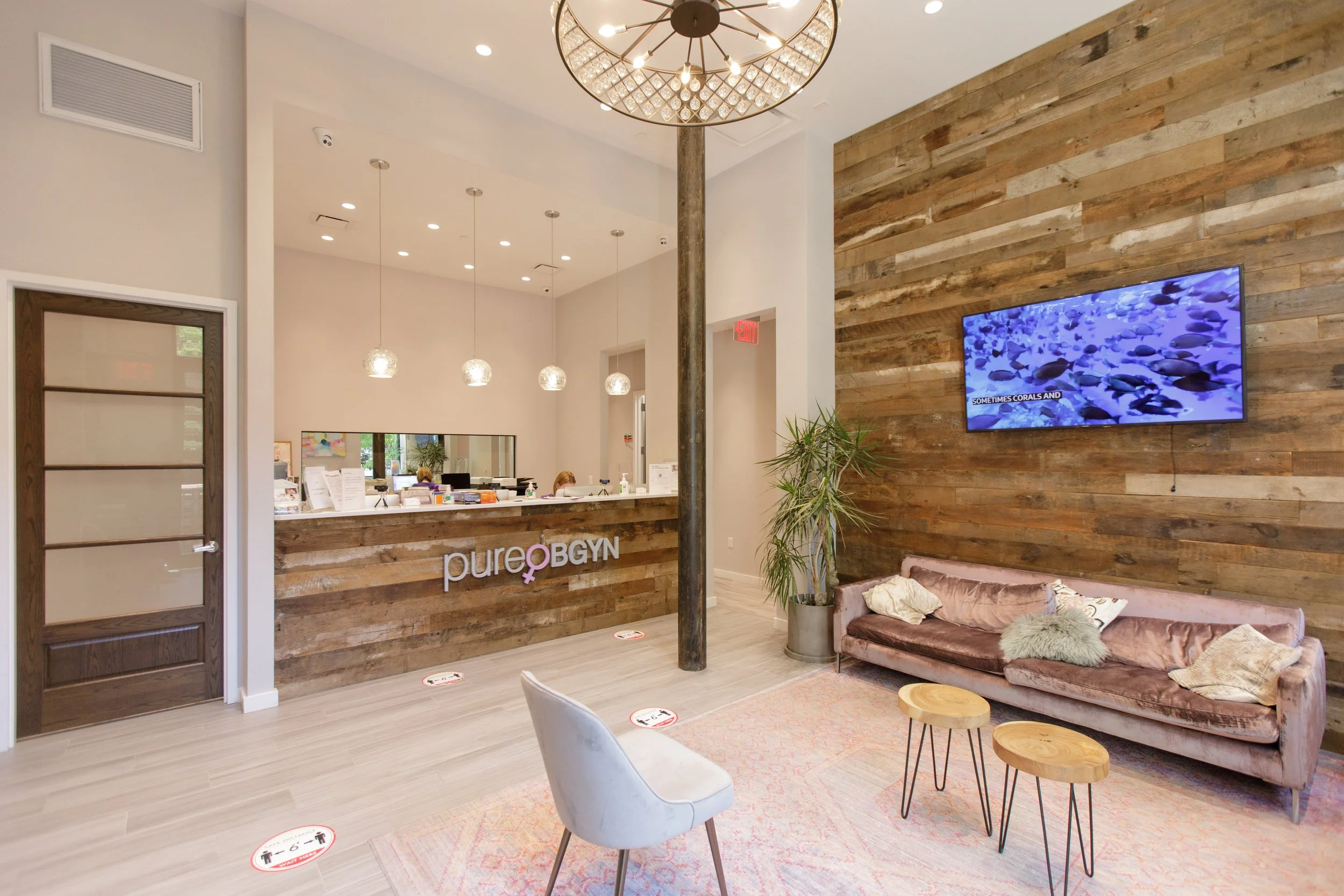 Reception area of a wellness or medical facility with a front desk, seating area with a pink velvet couch and small round wooden tables, a wall-mounted television, and a wooden accent wall. There are social distancing markers on the floor, a plant ne