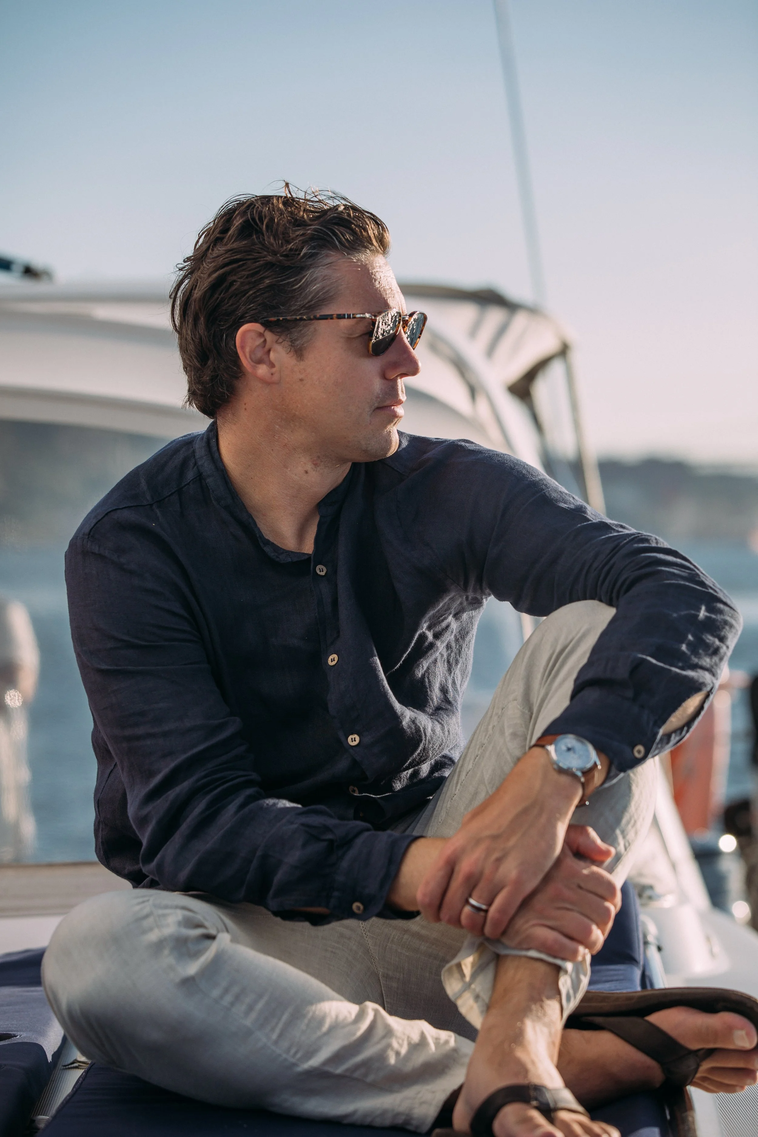 A man with brown hair and sunglasses sitting on a boat, looking to the side, with a calm water background.
