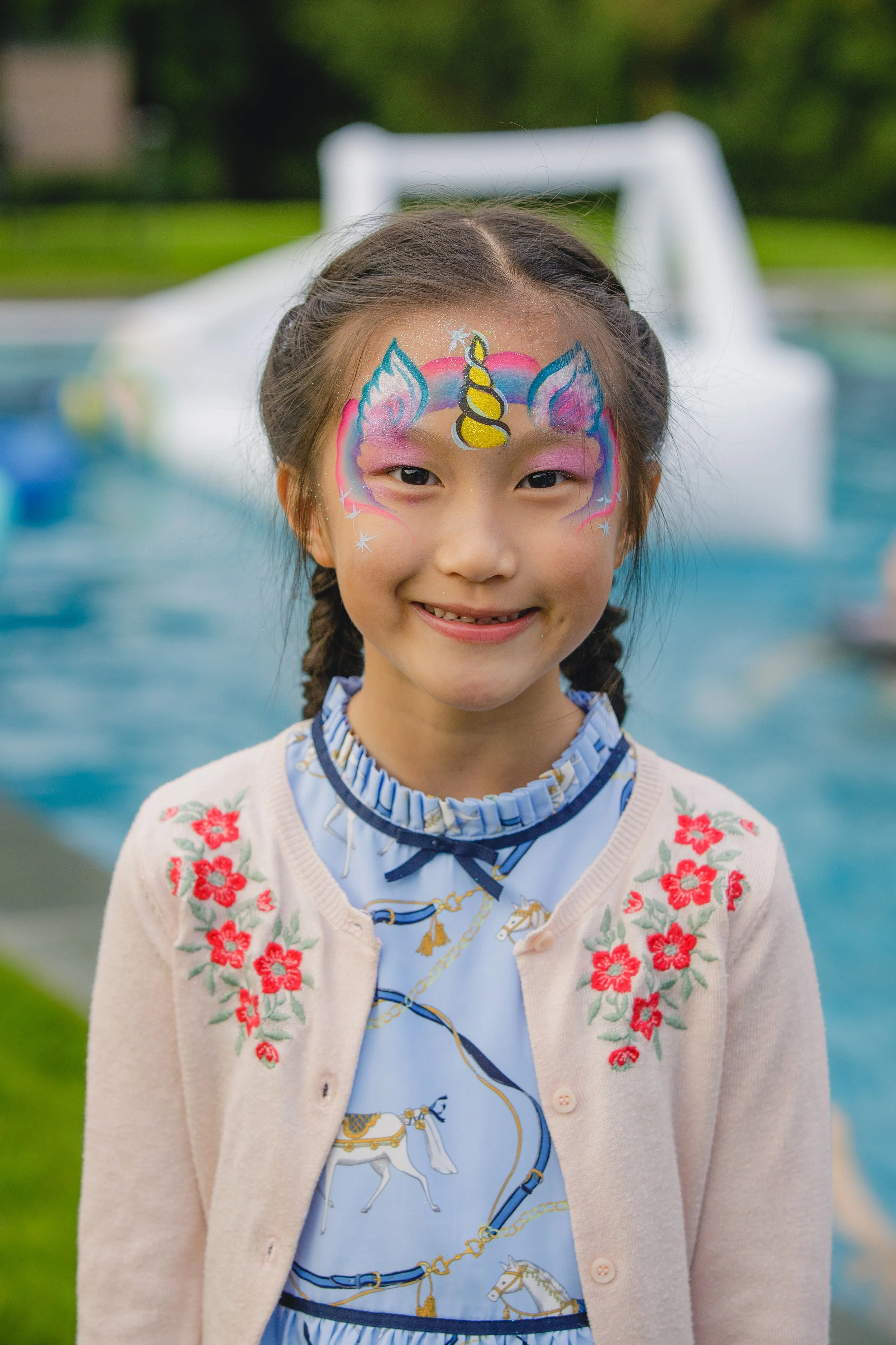 Young girl with braided hair smiling at the camera, face painted as a colorful unicorn with blue, pink, and yellow accents, standing outdoors near a pool with a white pool slide in the background.