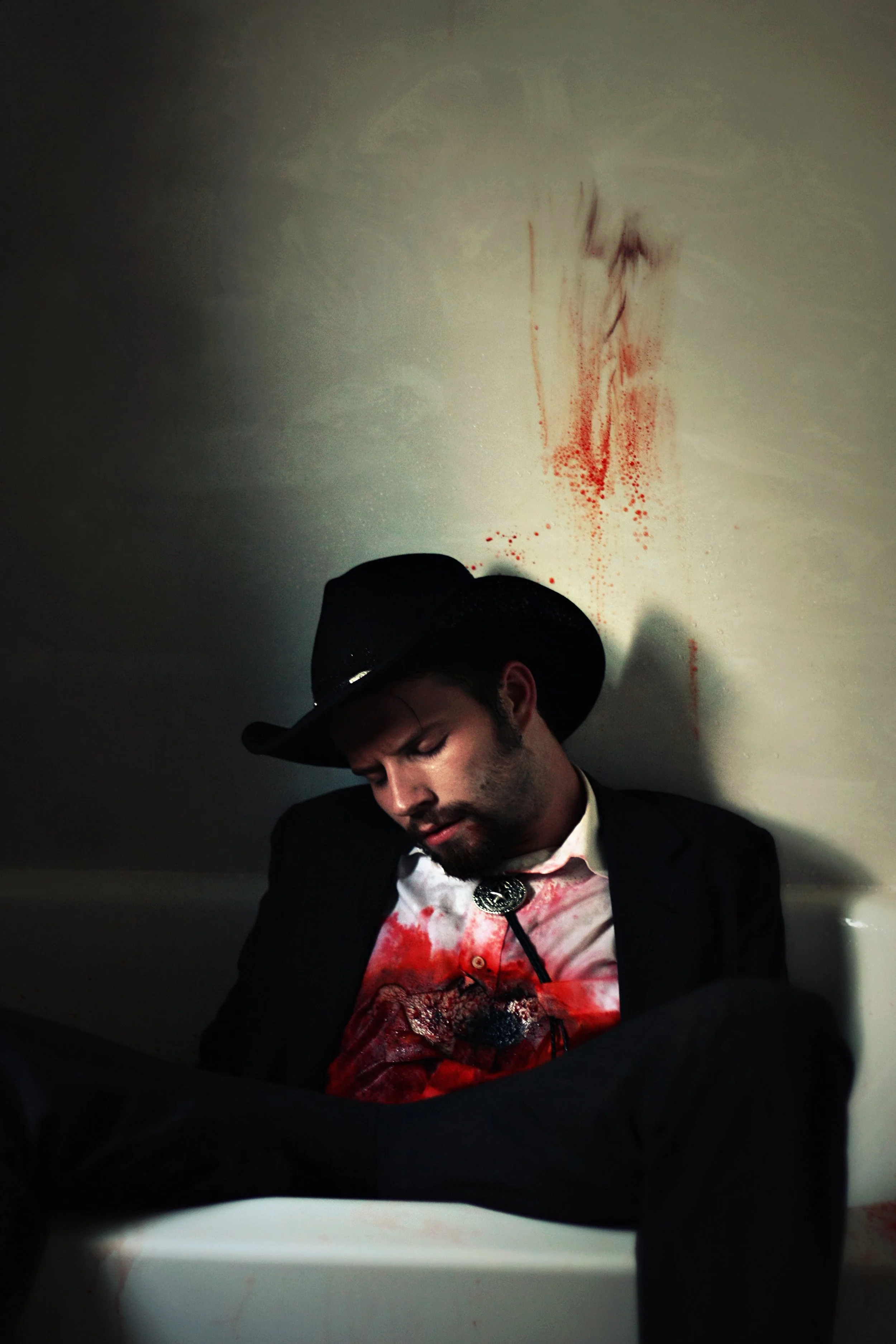 A man in a suit and cowboy hat with bloodstains on his shirt, sitting with his head down, in front of a white wall with blood splatters.