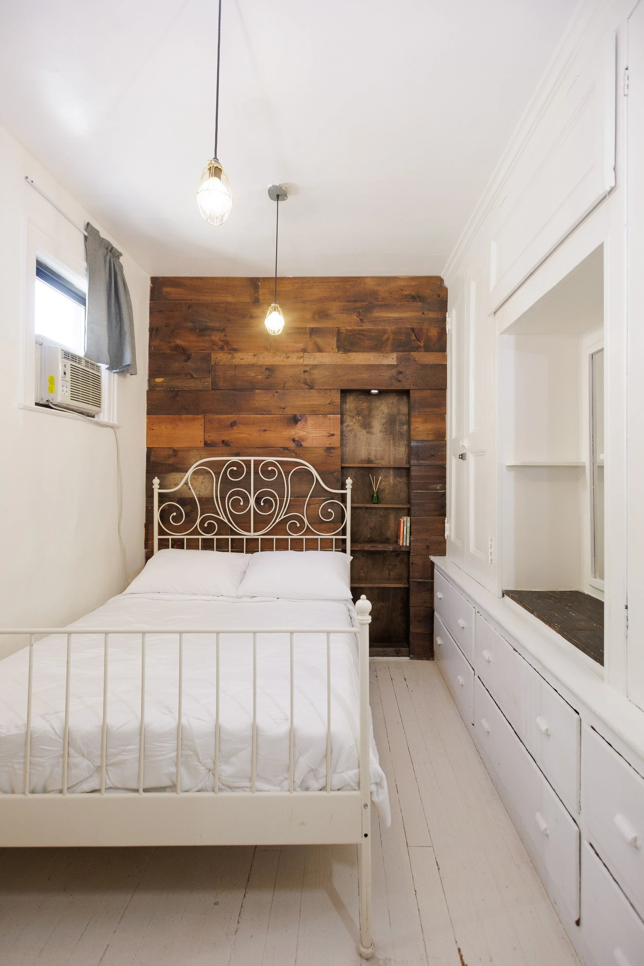 A bedroom featuring a white metal bed frame with a white mattress, a wood-paneled accent wall behind the bed, two hanging pendant lights, a small built-in shelf with a small plant and books, an air conditioning unit in the window, white walls and har