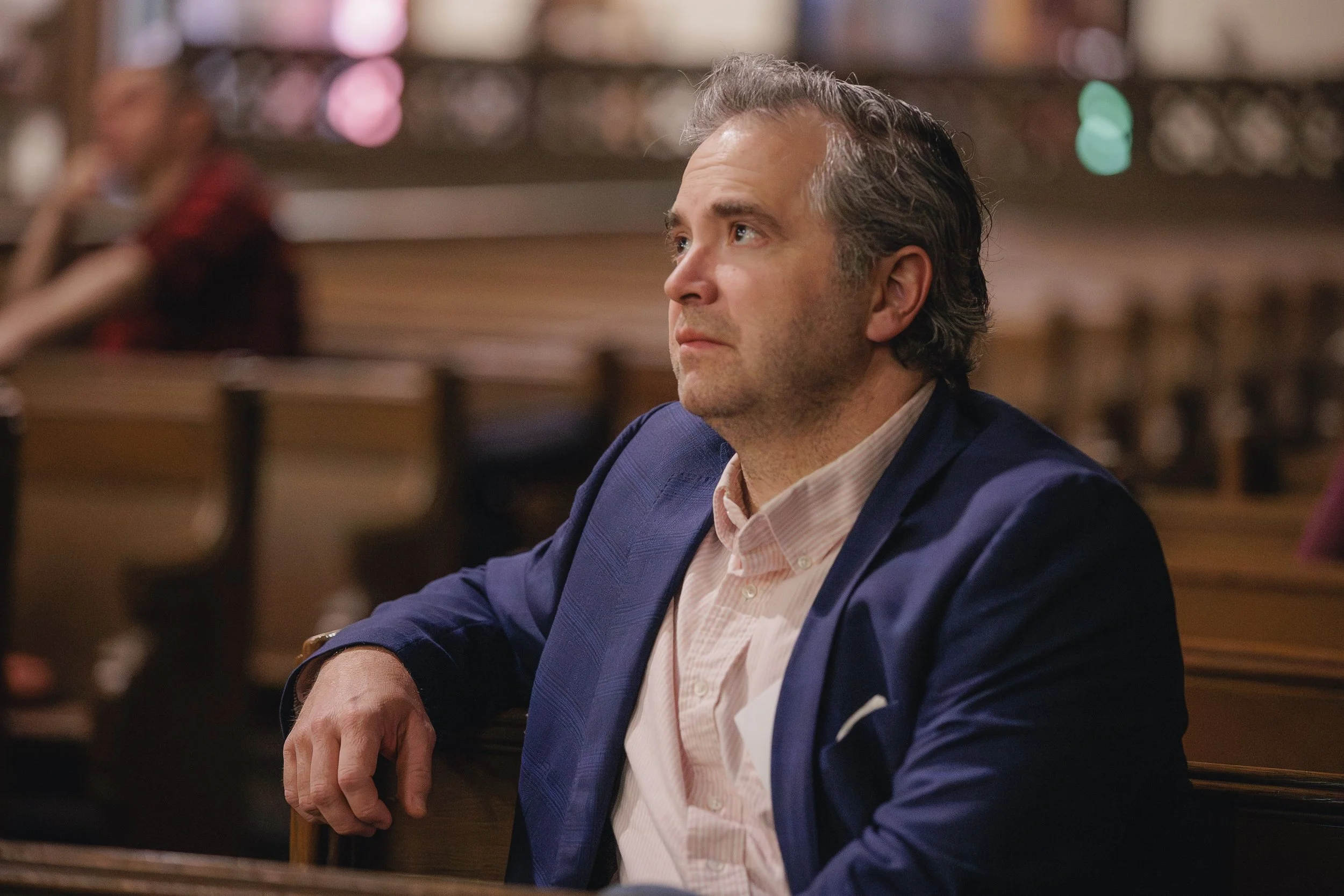 A man with wavy gray hair, wearing a blue blazer and a light pink shirt, sitting at a table in a restaurant with a blurred background of lights and shelves.
