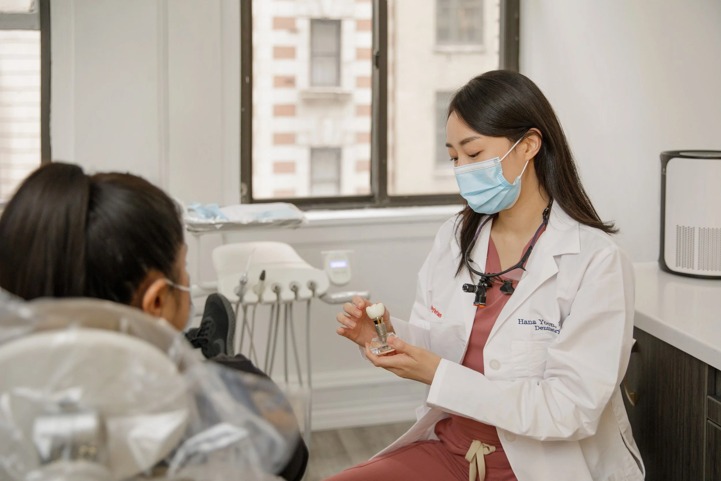 A female dentist wearing a face mask and dental loupes shows a dental implant to a patient in a dental office.