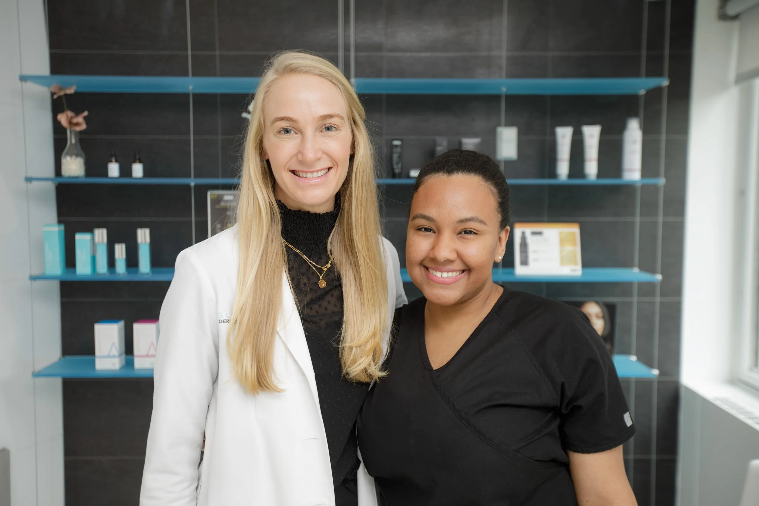 Two women smiling, one with long blonde hair wearing a white coat and the other with short dark hair wearing medical scrubs, standing in a professional setting with shelves of skincare or medical products in the background.