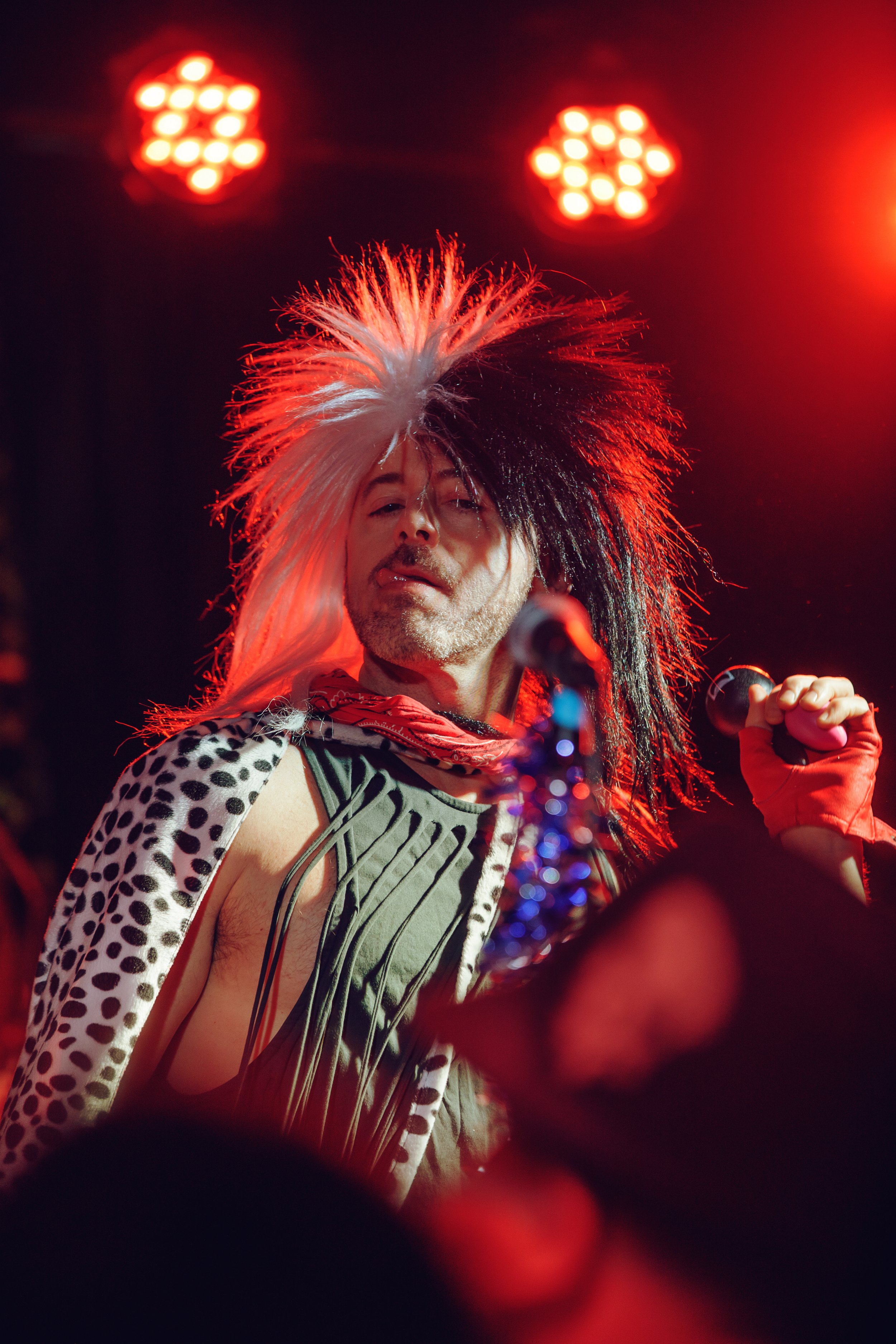Person performing on stage with a large, spiky, black and white wig, wearing a leopard print and striped outfit, holding a microphone, illuminated by red stage lights.
