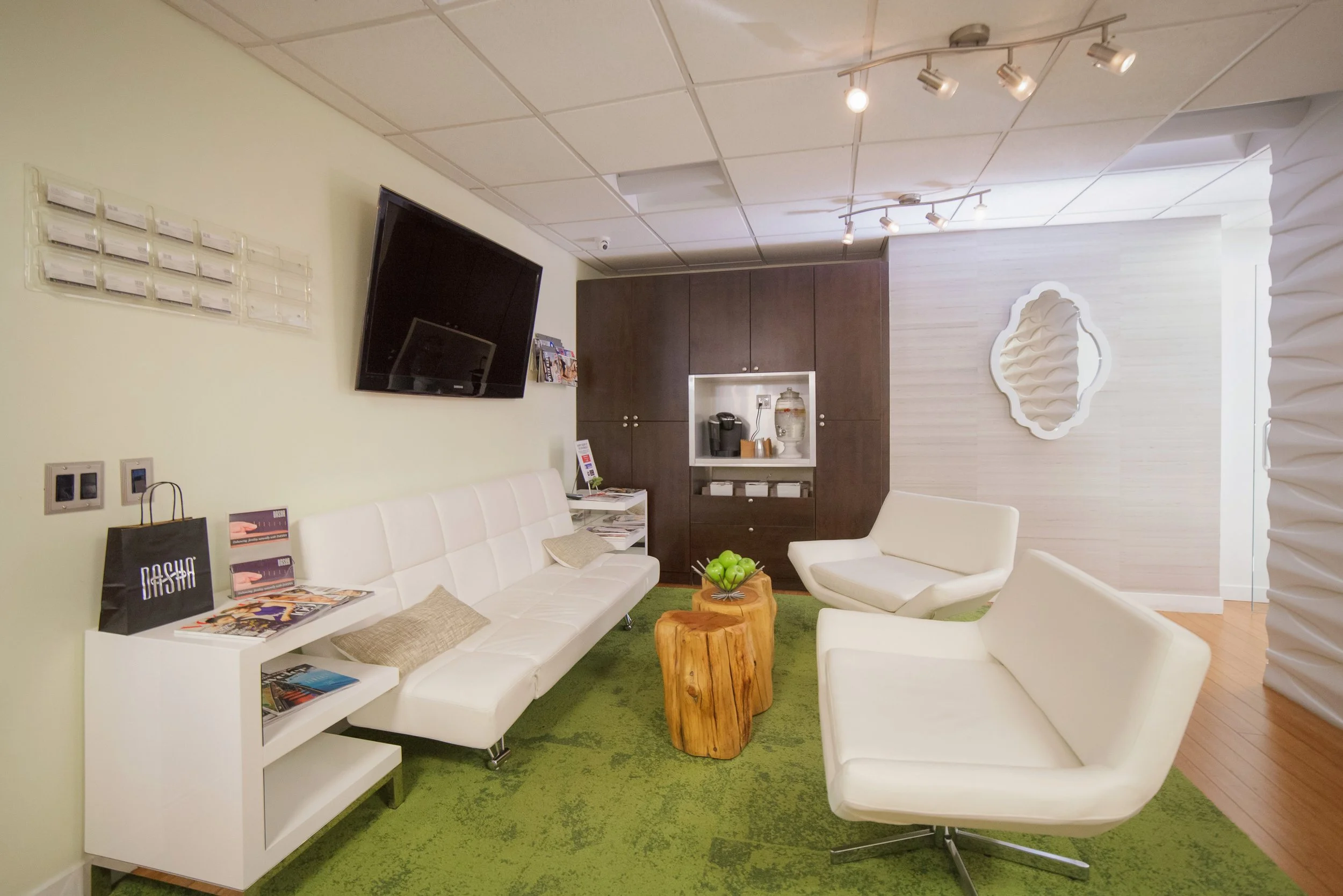 A cozy waiting room or lounge area with white chairs, a white sofa, and a small wooden side table with green apples. There is a large dark wood cabinet, a wall-mounted flat-screen TV, and a decorative white mirror on the light-colored wall. The ceili