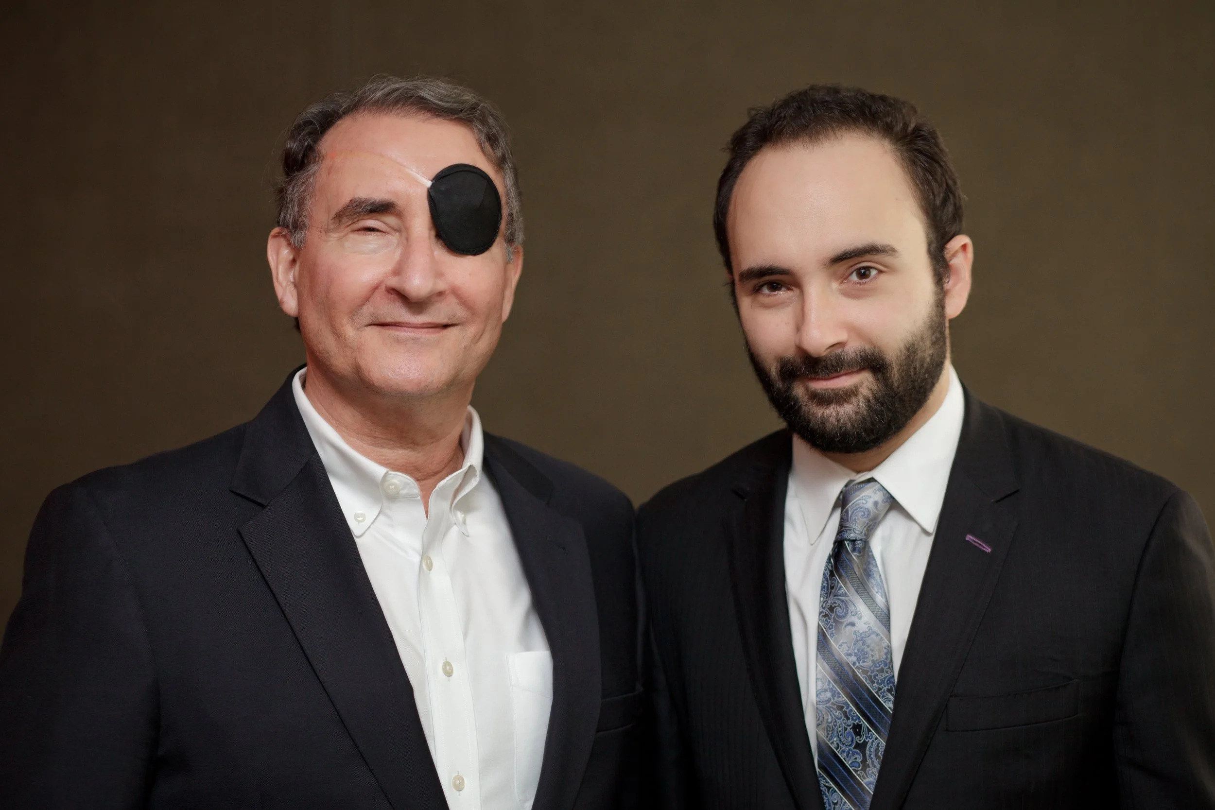 Two men in formal suits standing side by side, smiling, with a plain brown background. The man on the left wears a black eye patch over his right eye, and the man on the right has a full beard.