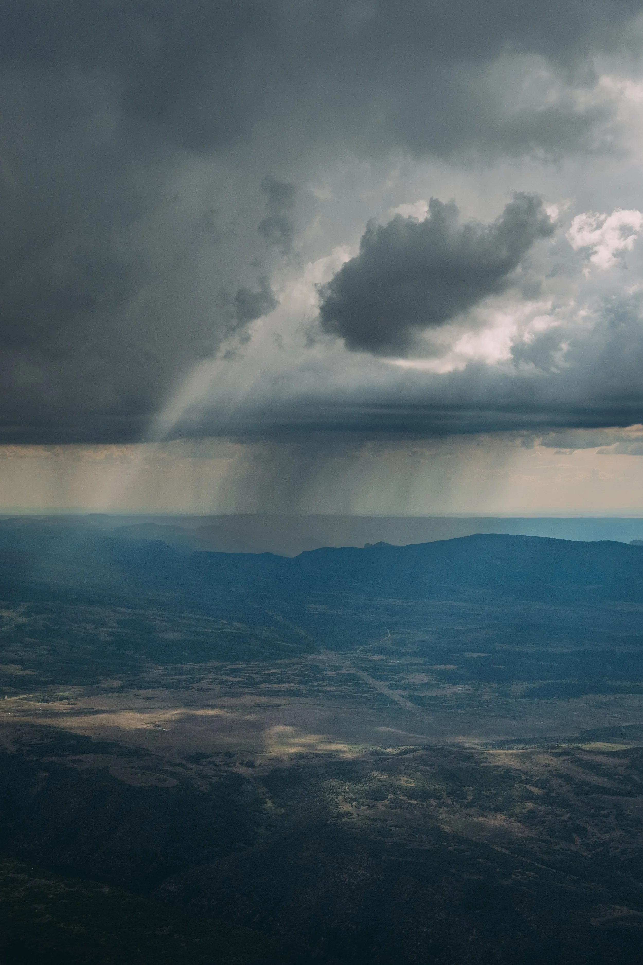A view of a cloudy sky with rainfall visible over a vast landscape with mountains and flat land below.