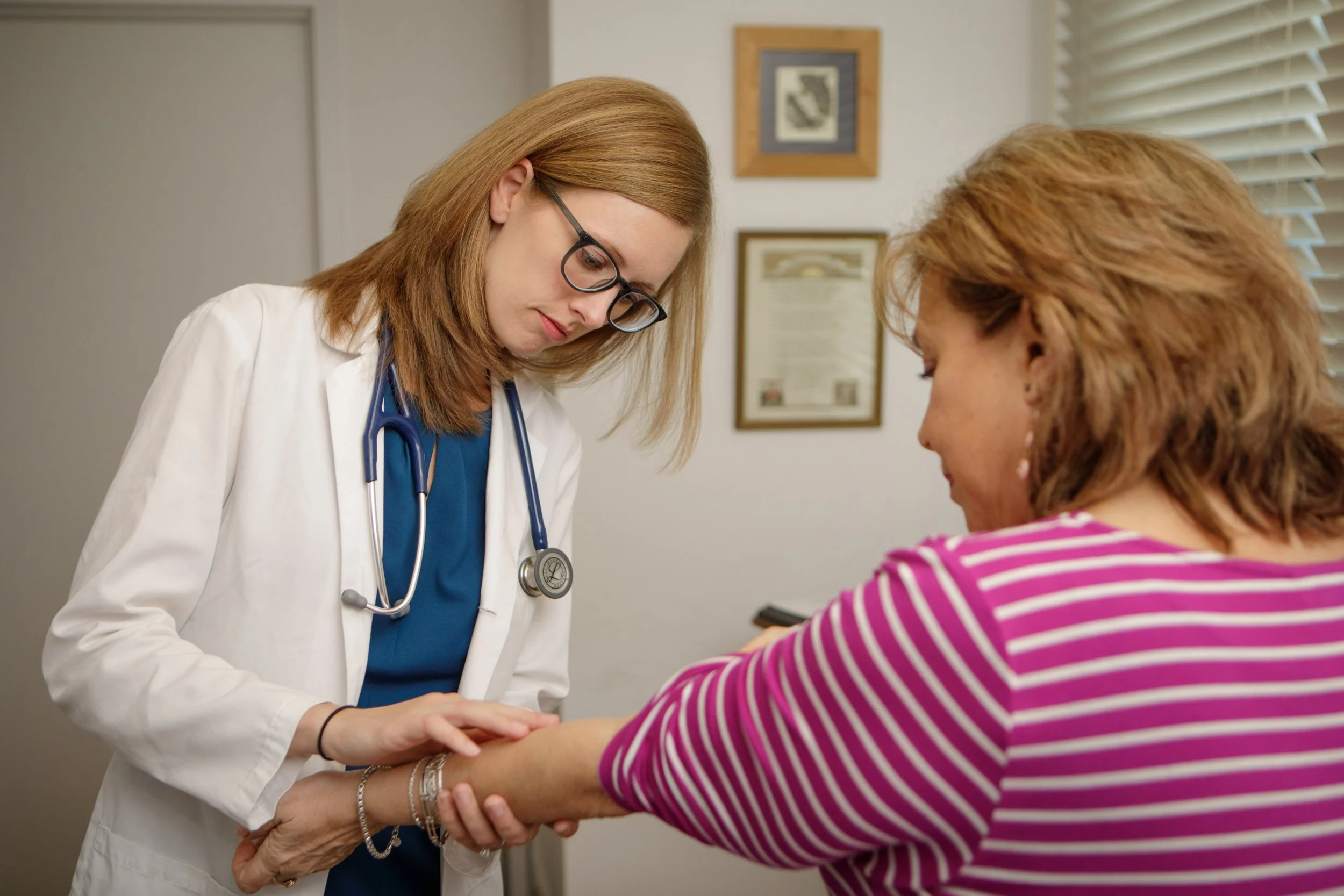 A healthcare professional, wearing glasses and a white coat with a stethoscope around her neck, gently holds a patient's hand and forearm during a consultation in a medical office.