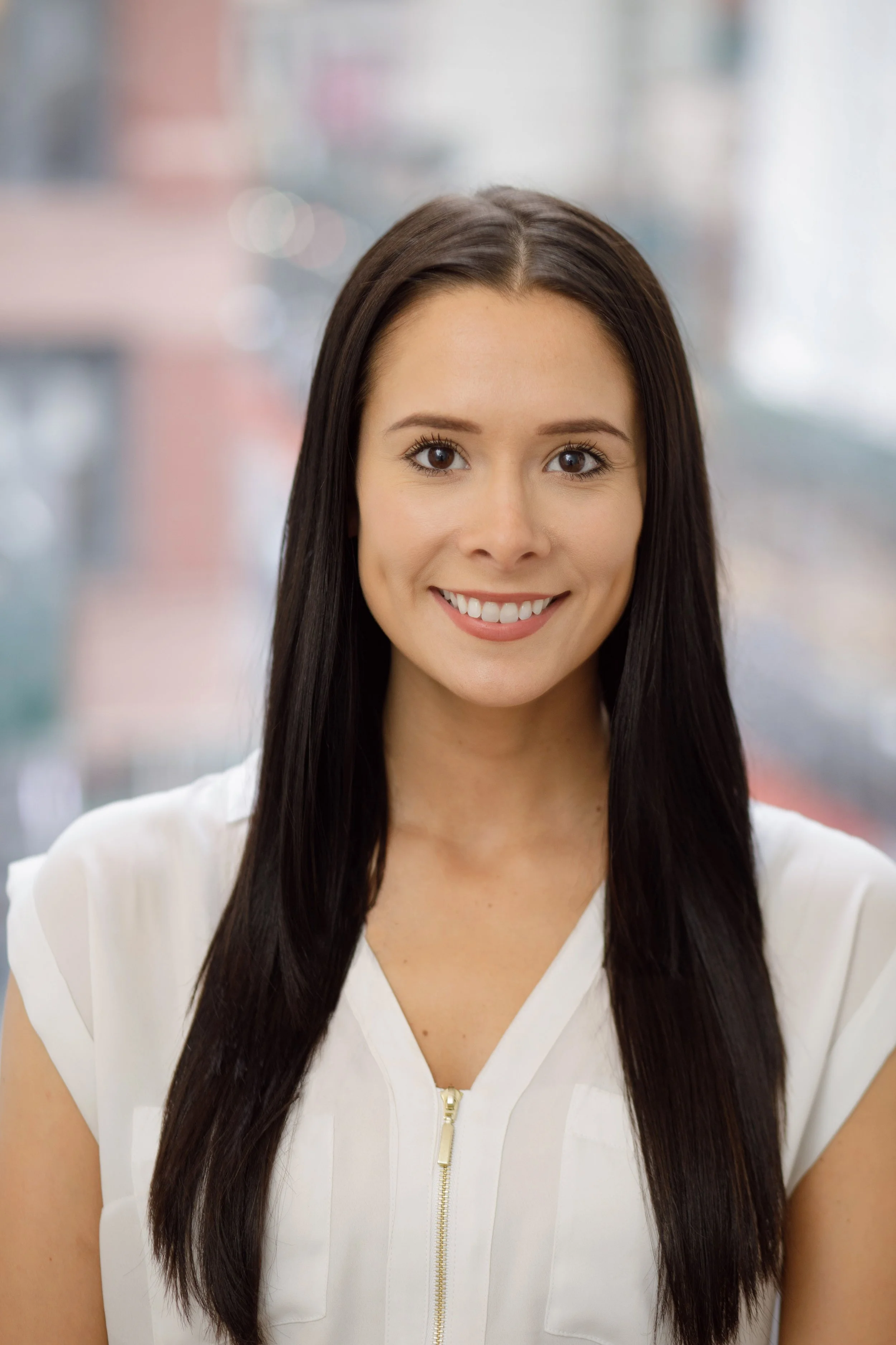 A young woman with straight dark brown hair and brown eyes, smiling, wearing a white top with a zipper, in a blurred indoor setting.