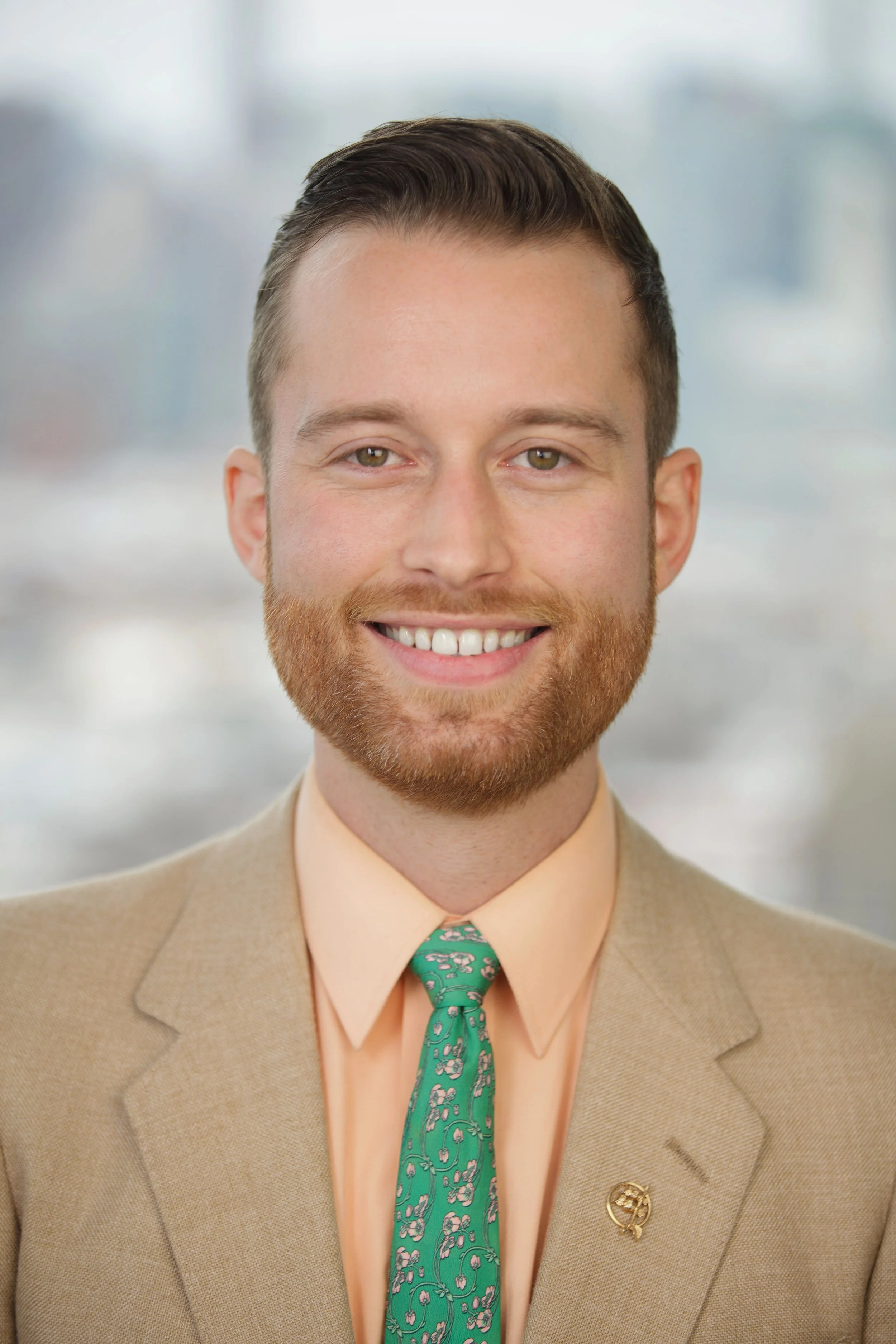A young man with light skin, brown hair, and a beard, smiling and wearing a beige suit, light pink shirt, and a green tie with white floral pattern.