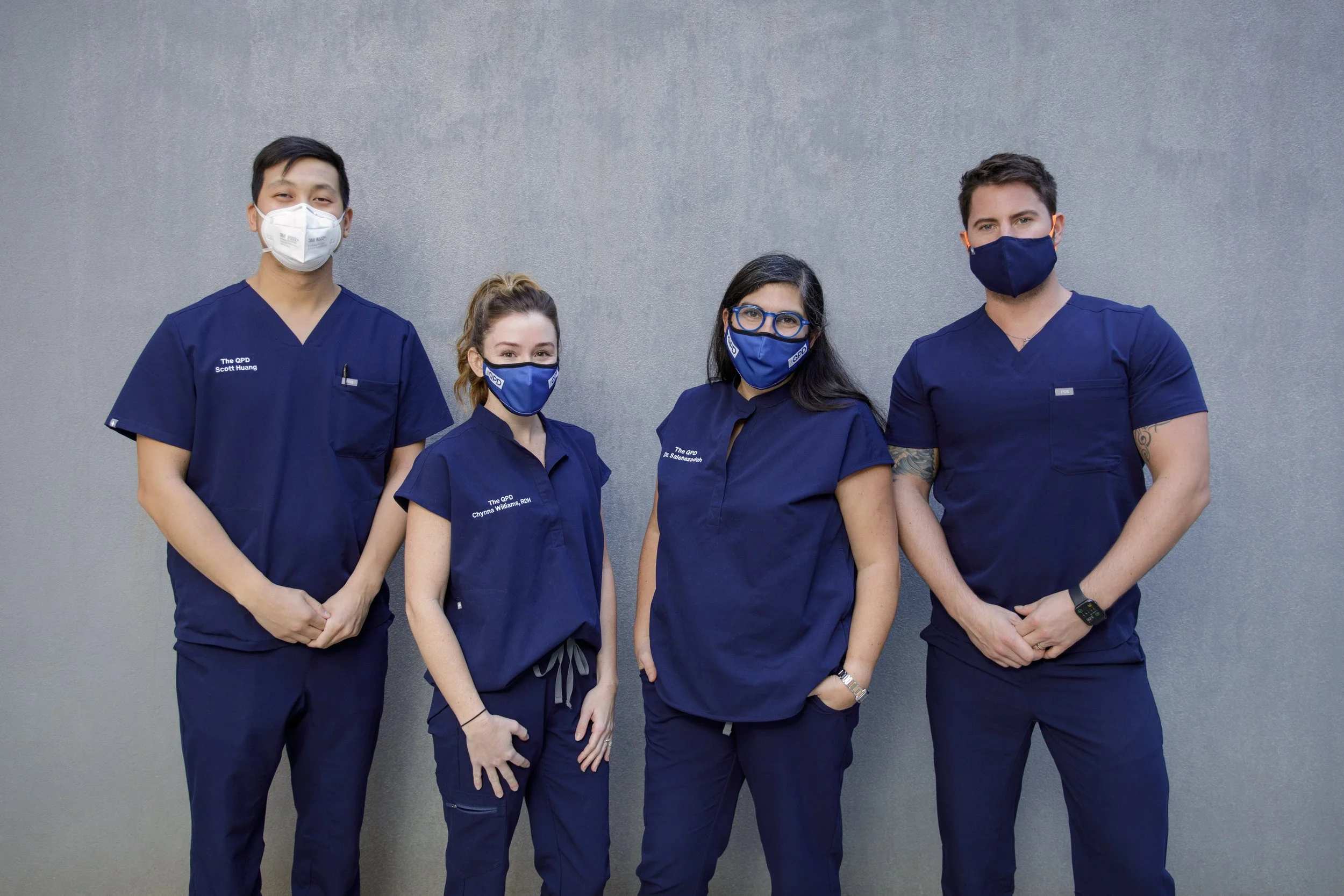 Four medical professionals standing against a plain gray wall, wearing blue scrubs and face masks. The two men are on the ends, and the women are in between. They are posing confidently for the photo.