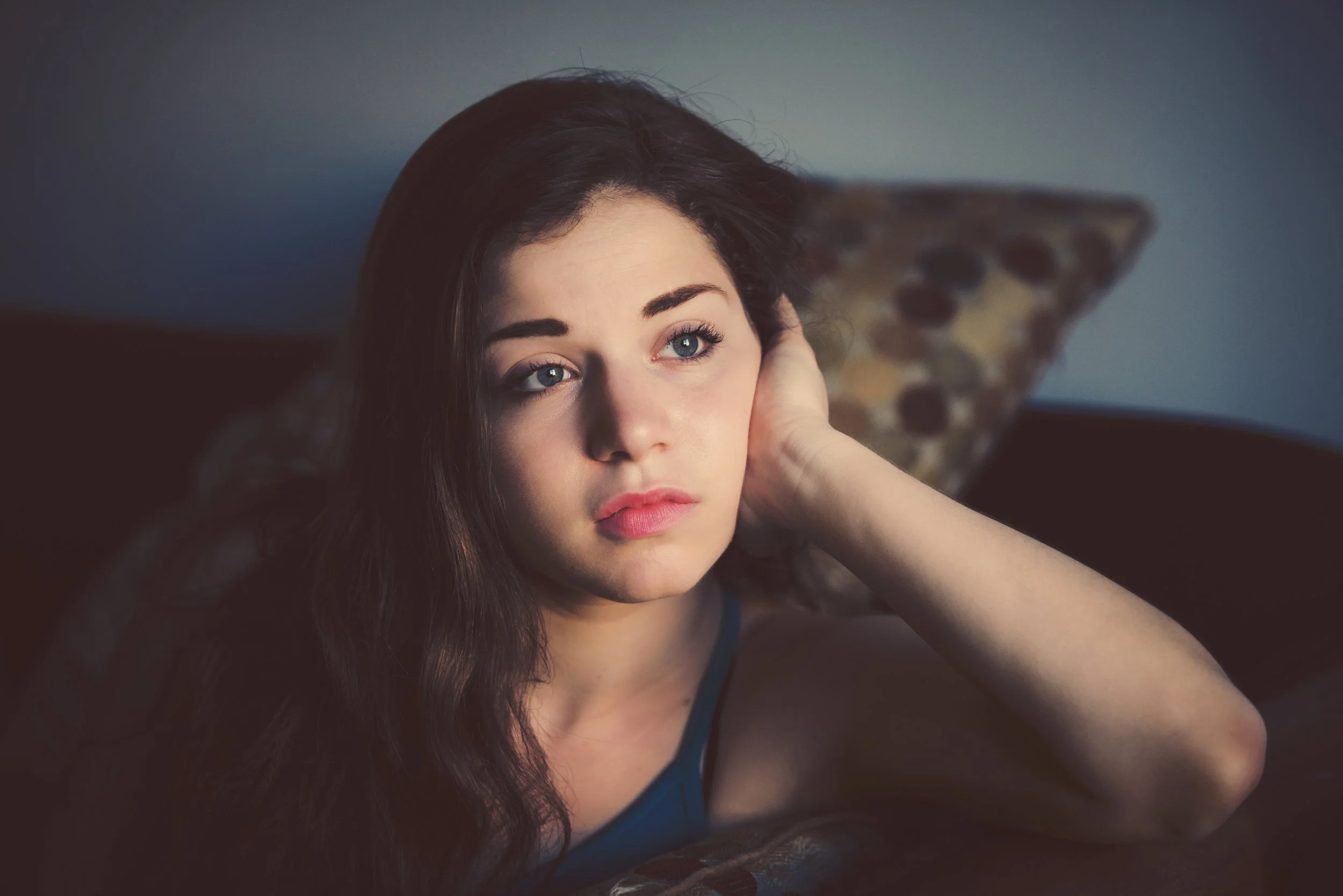 A young woman with blue eyes and long brown hair relaxing on a couch with a patterned pillow behind her.