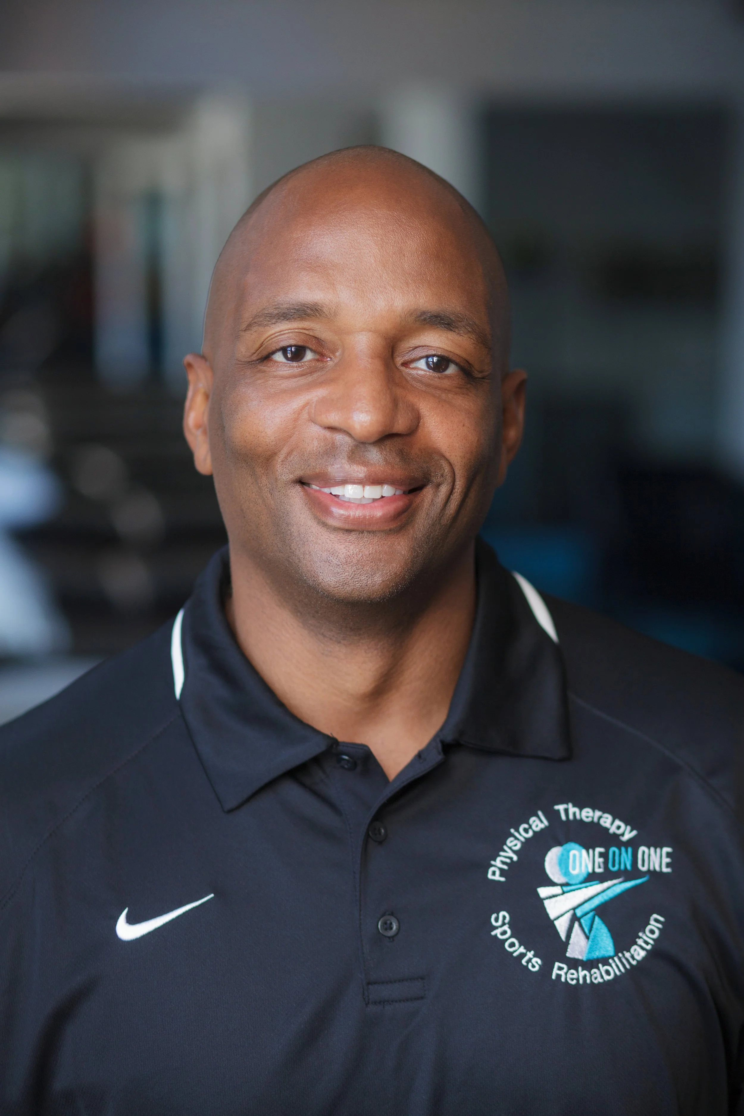 Close-up of a smiling African American man wearing a black polo shirt with a logo for Physical Therapy Sports Rehabilitation on the chest. The background is blurred.