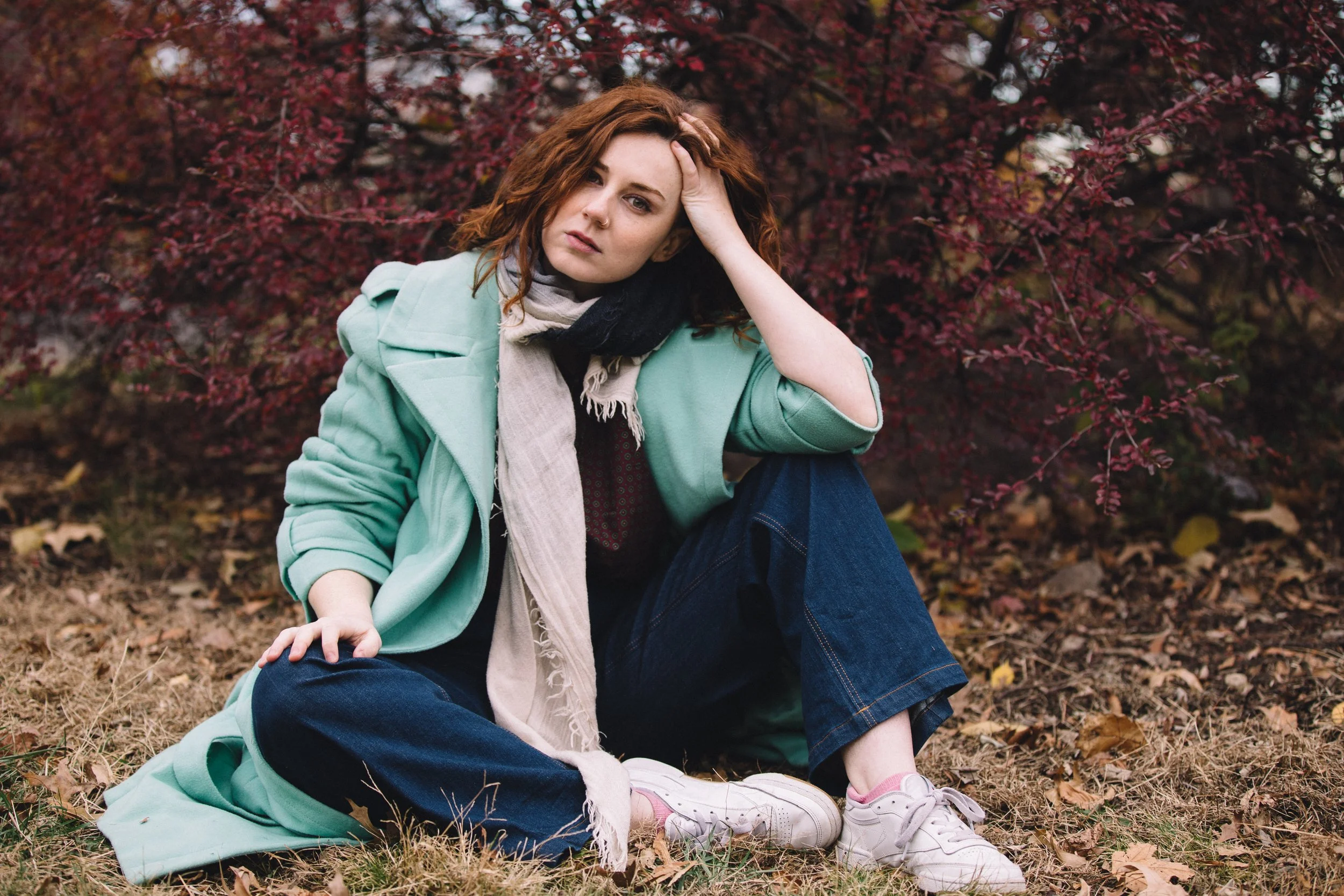 A young woman with curly red hair and a nose piercing sitting on fallen leaves in front of a dark red bush, wearing a mint green coat, a beige scarf, and white sneakers, with her hand on her forehead, looking thoughtful.