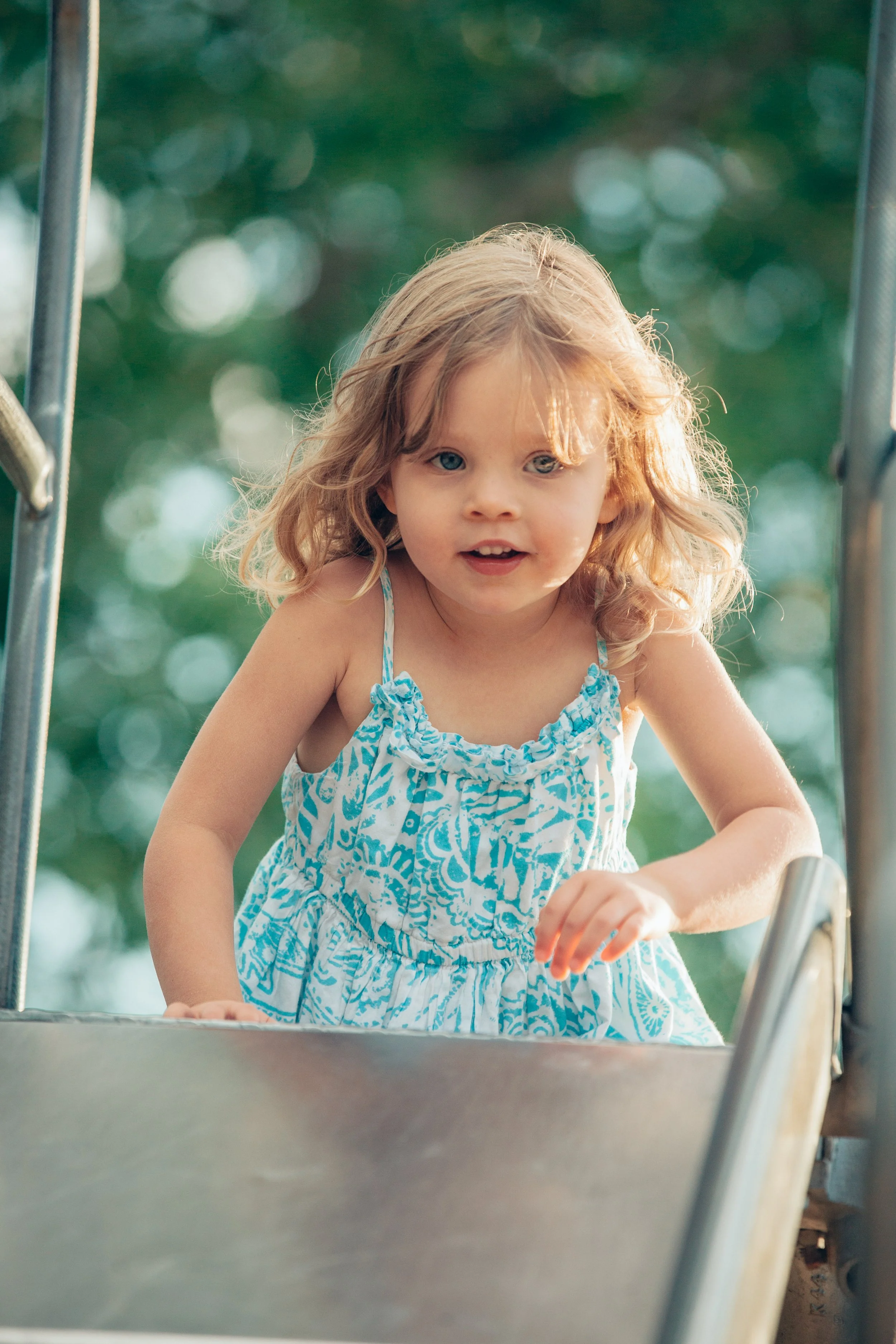 A young girl with curly blonde hair and blue eyes wearing a blue and white patterned dress, standing at the top of a slide outdoors, looking at the camera with a curious expression. Blurred green trees are in the background.