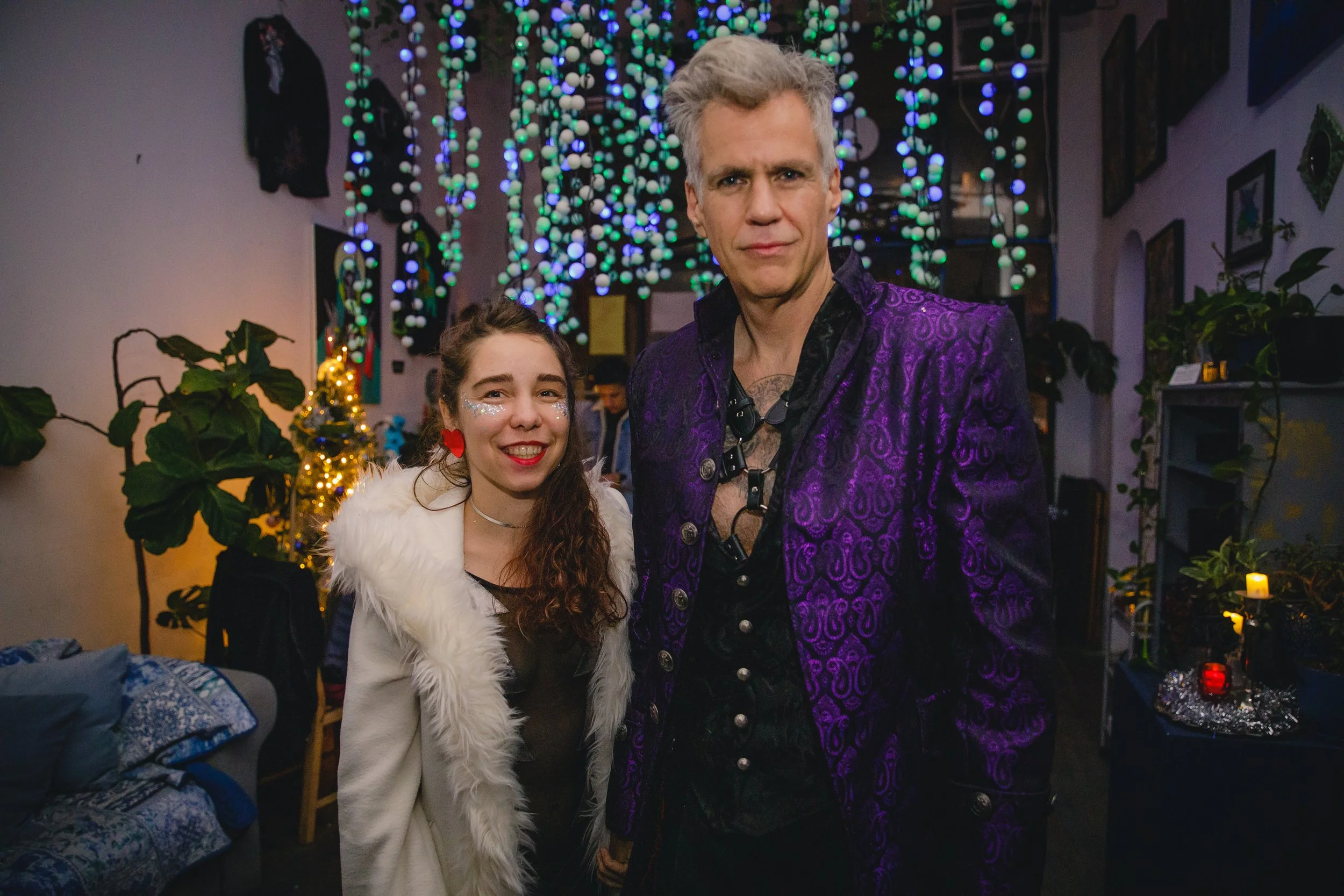 A young woman with curly hair, face glitter, red heart earrings, and a white furry jacket wrinkles her nose and smiles, standing next to a tall man with gray hair in a purple patterned blazer in a festive room with colorful hanging lights, green plan