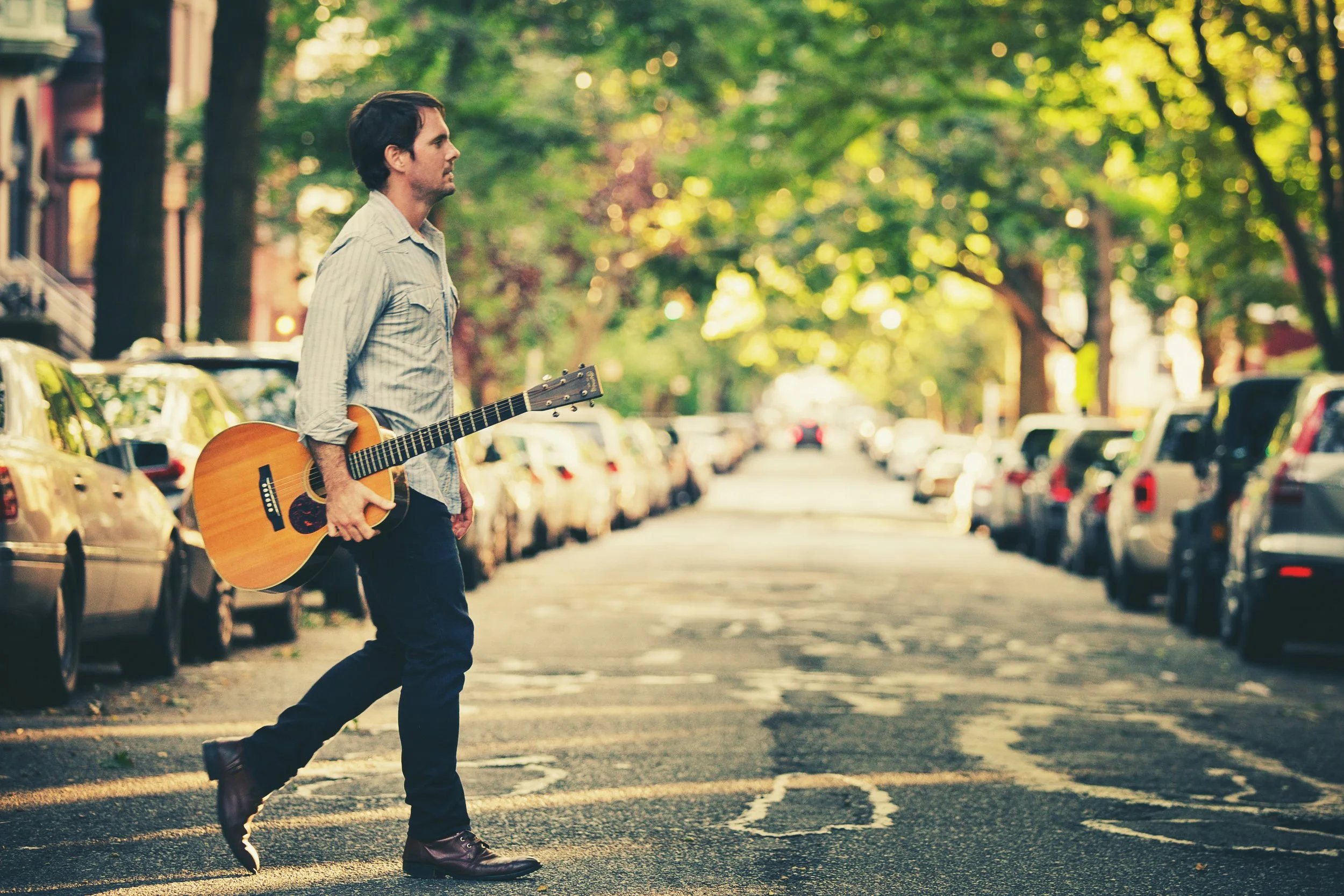 A man walking across a city street with a guitar, with cars parked along the street and trees with green leaves in the background.
